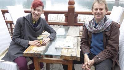 Michael Wood sitting at a table by a lake with a young stylishly-dressed Chinese man