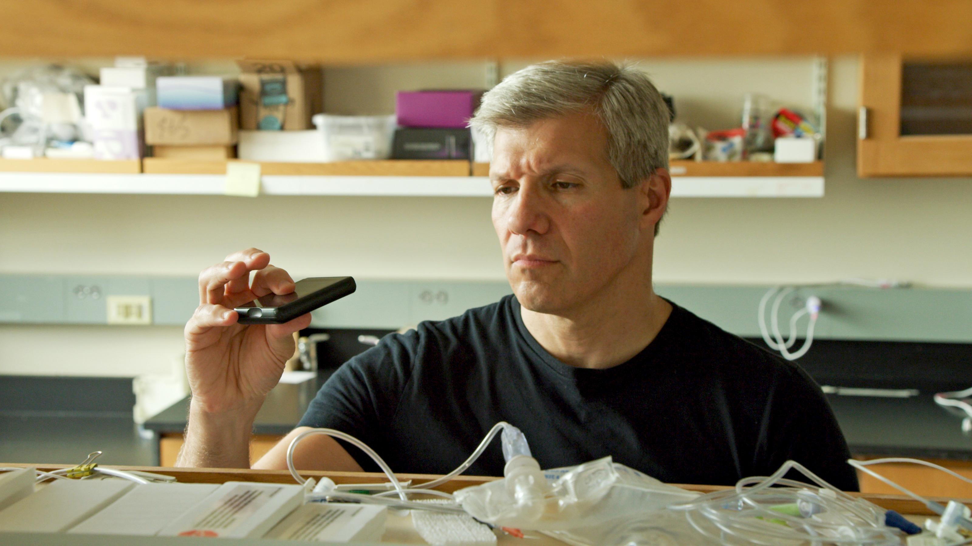 Ed Damiano holds up a black object about the size of a cell phone. before him is a shelf of medical supplies