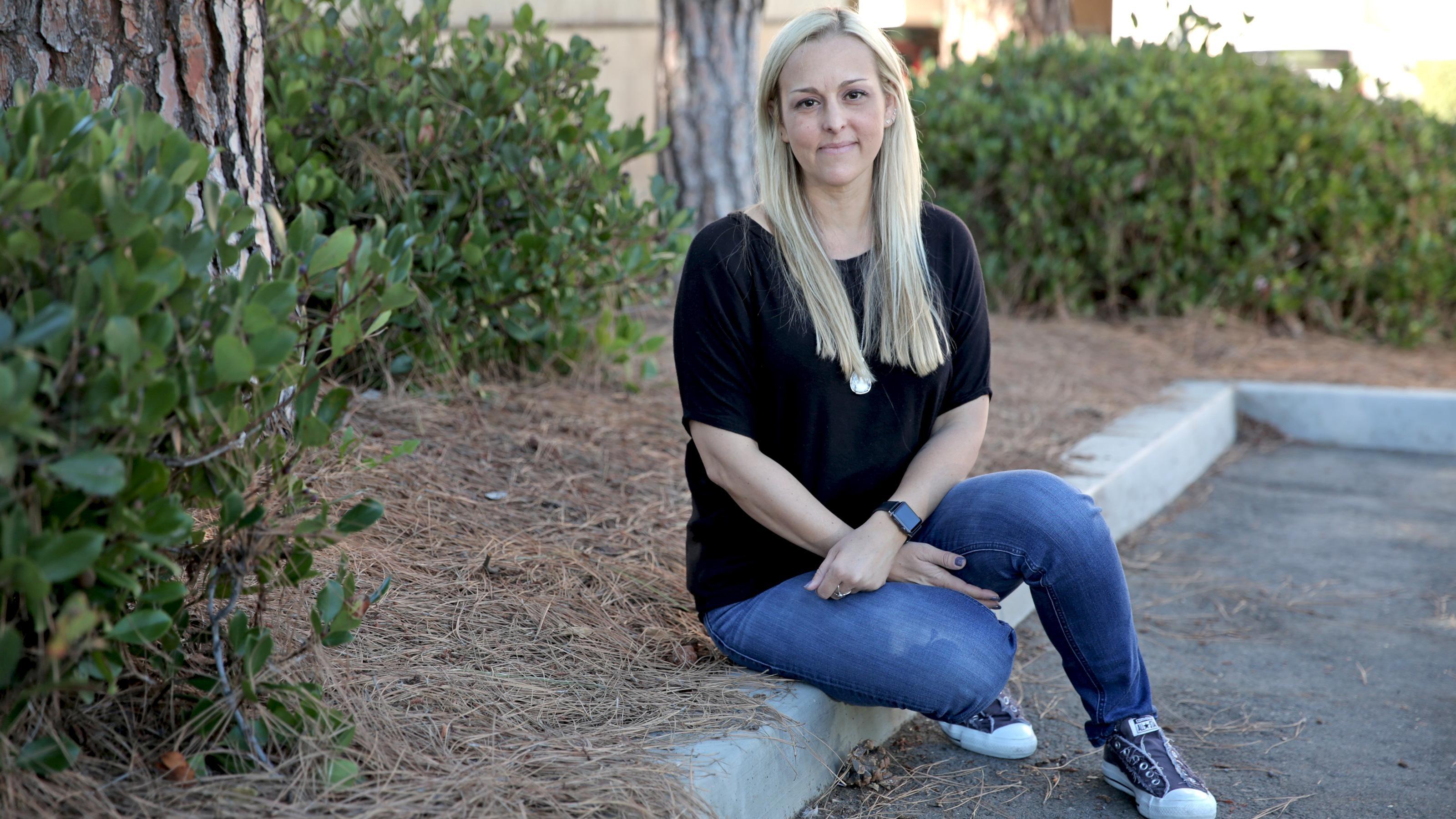 Nicole, wearing a black tee-shirt and jeans, sits on a curb.