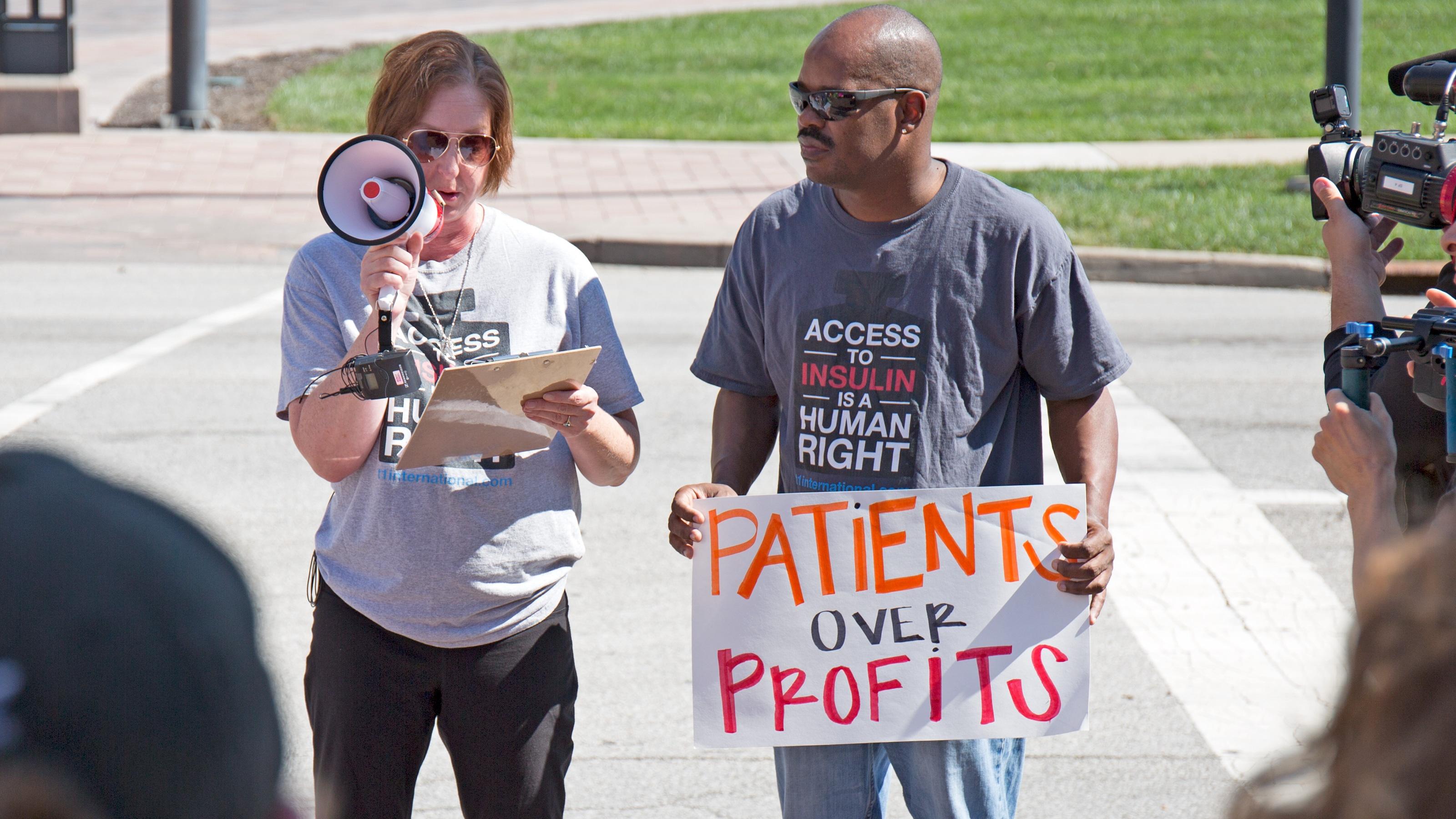 Nicole Smith-Holt holds a loud speaker as she protests outside a drug company about the high cost of insulin. Her husband stands beside her, holding a sign that reads: "Patients Over Profits." 