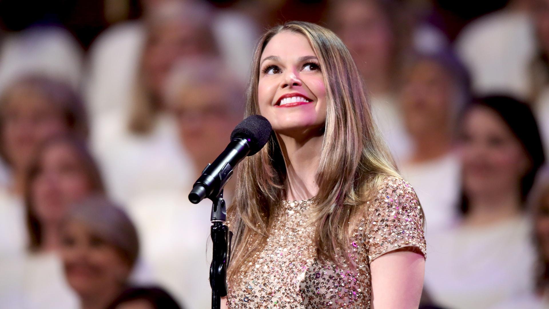 Kristin Chenoweth smiles on stage in front of a microphone with violinists playing behind her.