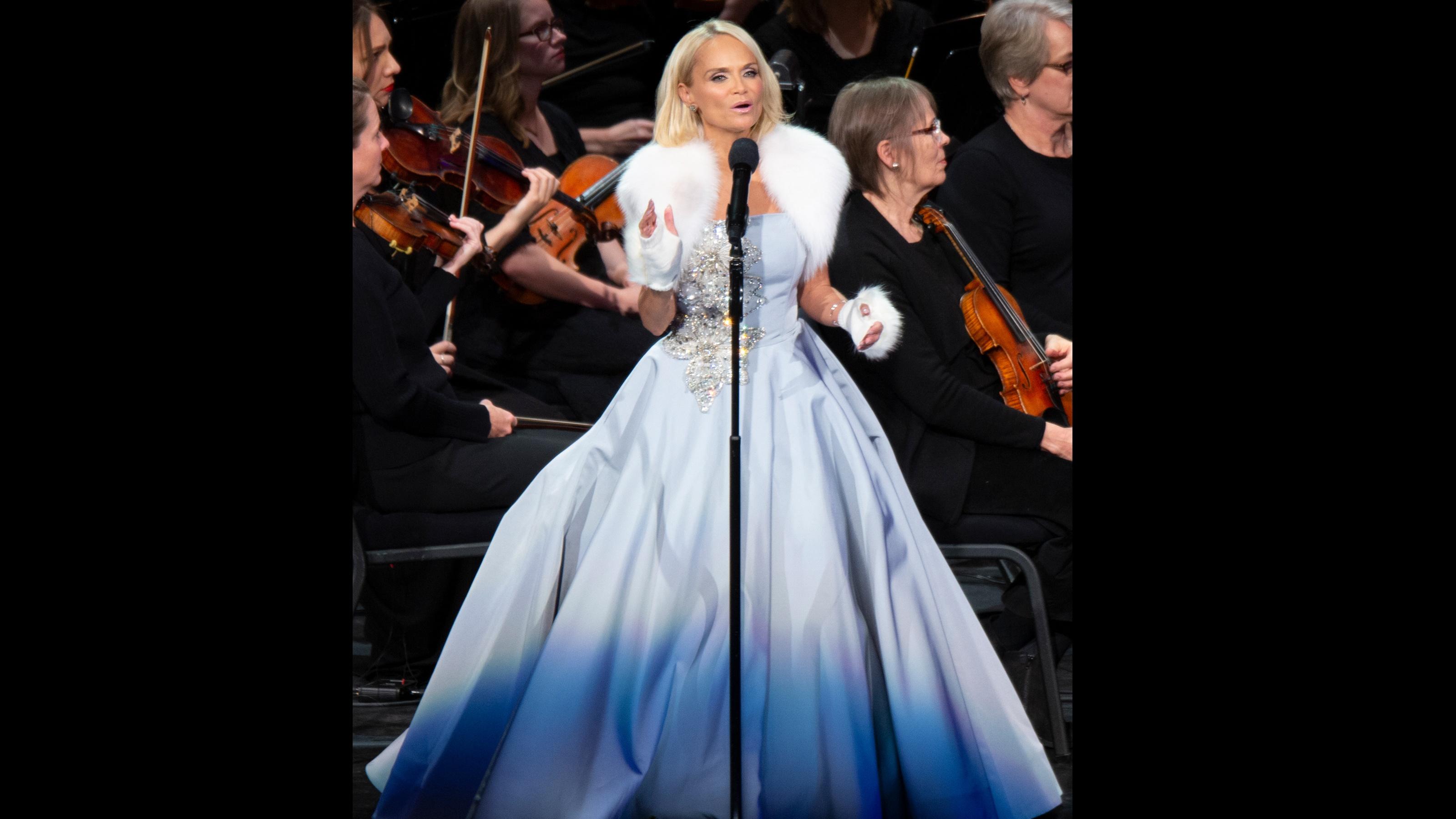 Kristin Chenoweth wears a pale blue dress with fur accents and sings in front of the violinists of The Orchestra at Temple Square.