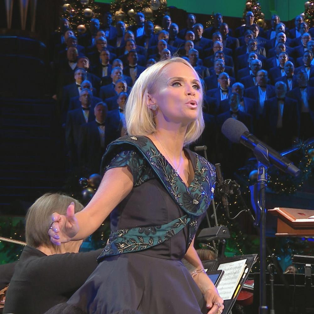 Kristin Chenoweth sings O Holy Night! into a microphone while The Tabernacle Choir accompanies behind her.