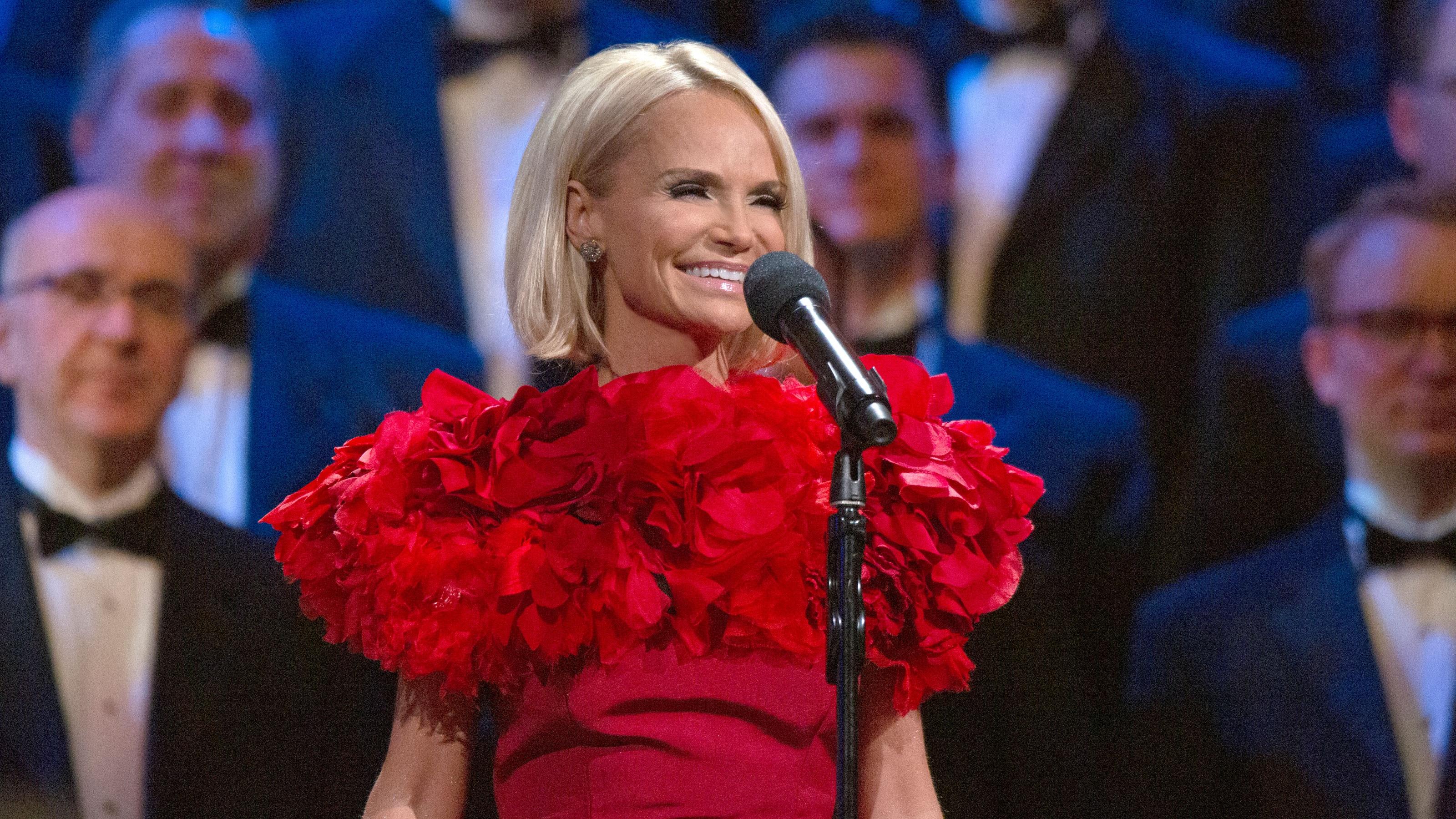 Kristin Chenoweth smiles in front of a microphone on a stand with The Tabernacle Choir behind her.