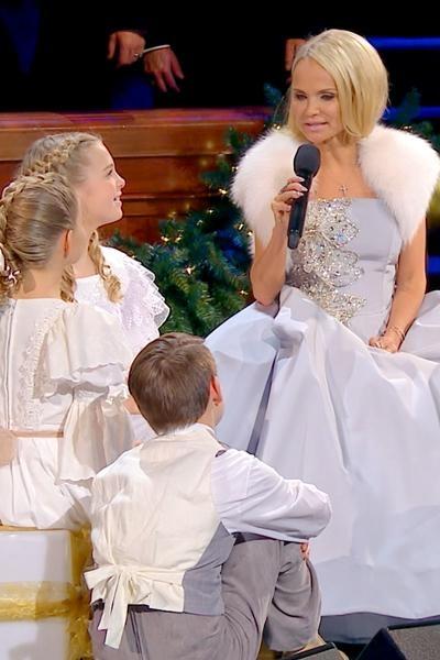 Sitting beside three children, Kristin Chenoweth sings What Child Is This? into a microphone.