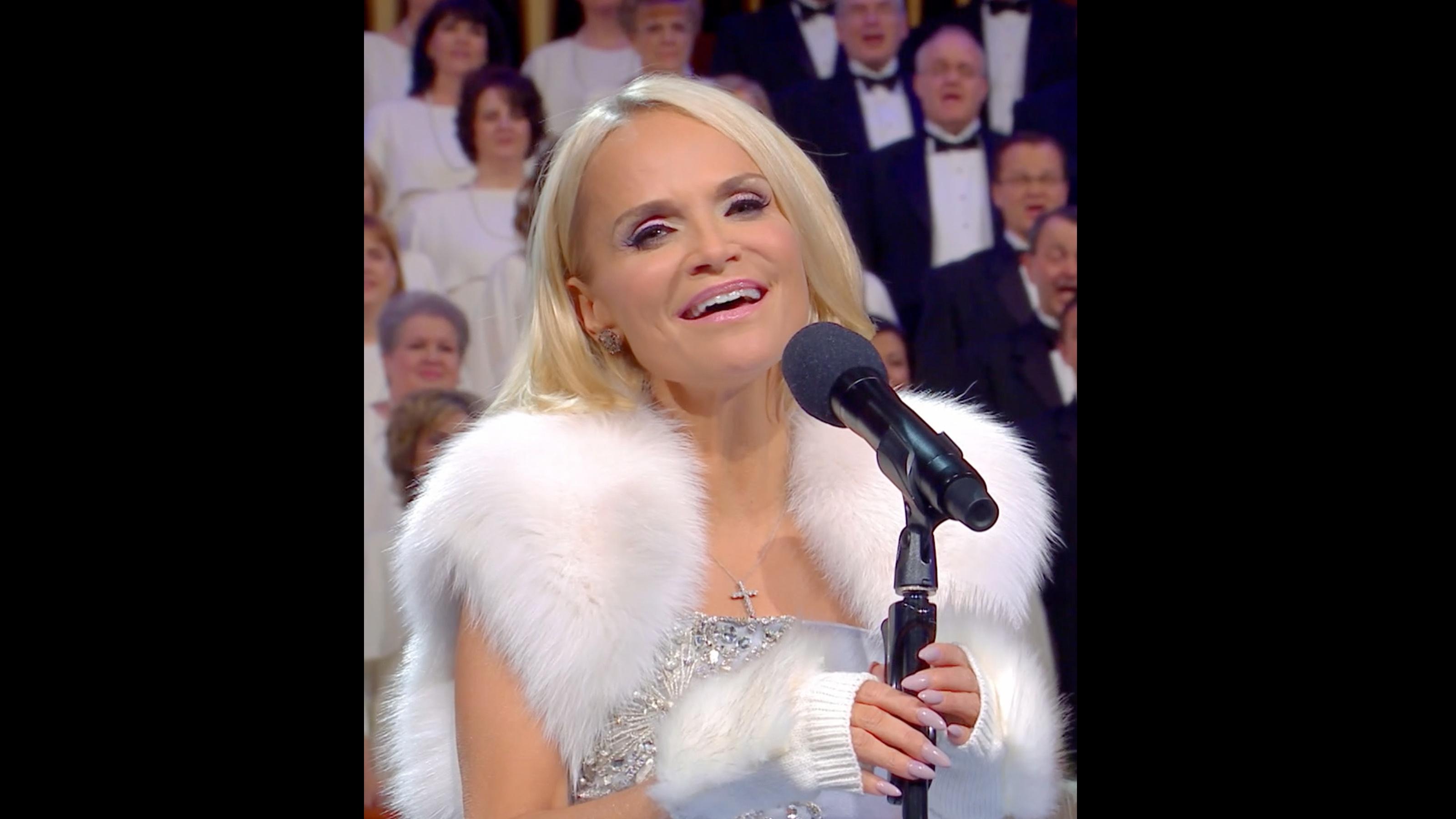 Kristin Chenoweth sings into a microphone on a stand while The Tabernacle Choir sings in the background.