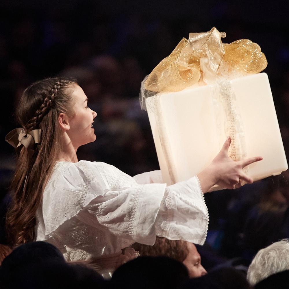 A young woman dressed in white holds up a wrapped, cream-colored present.