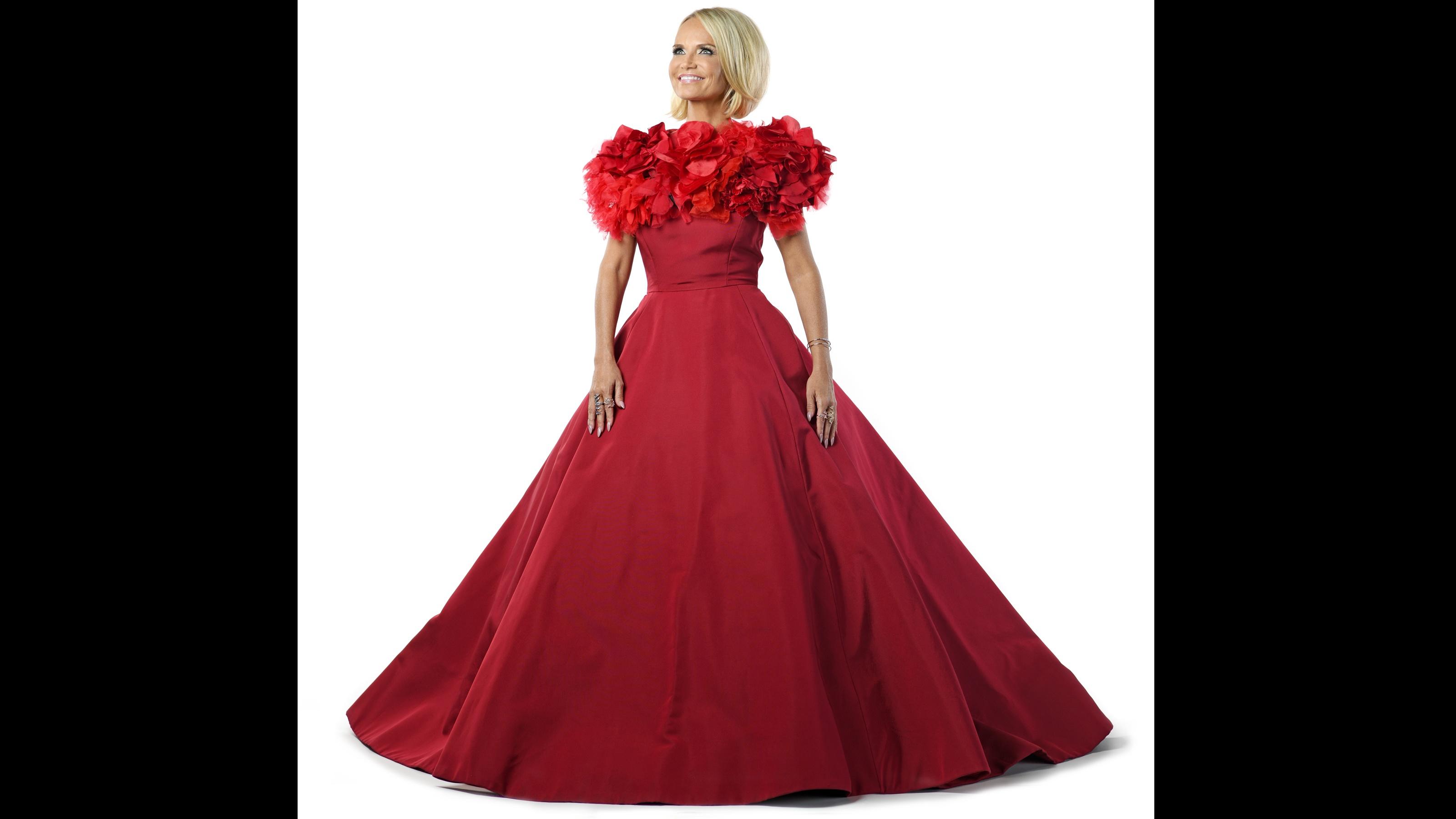 Kristin Chenoweth stands, smiling, in a red dress in front of a white background.