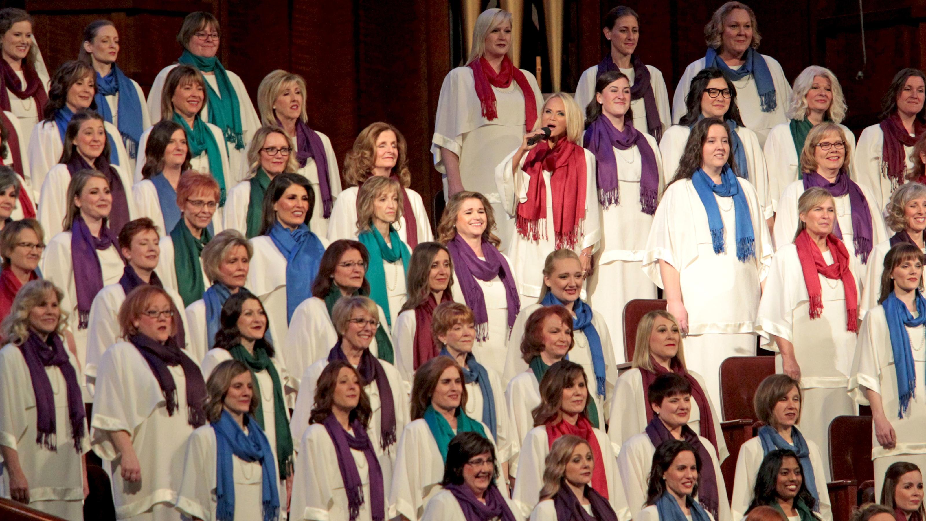 Kristin Chenoweth sings into a microphone amidst the female members of The Tabernacle Choir, who are all dressed in white with single-color scarves.