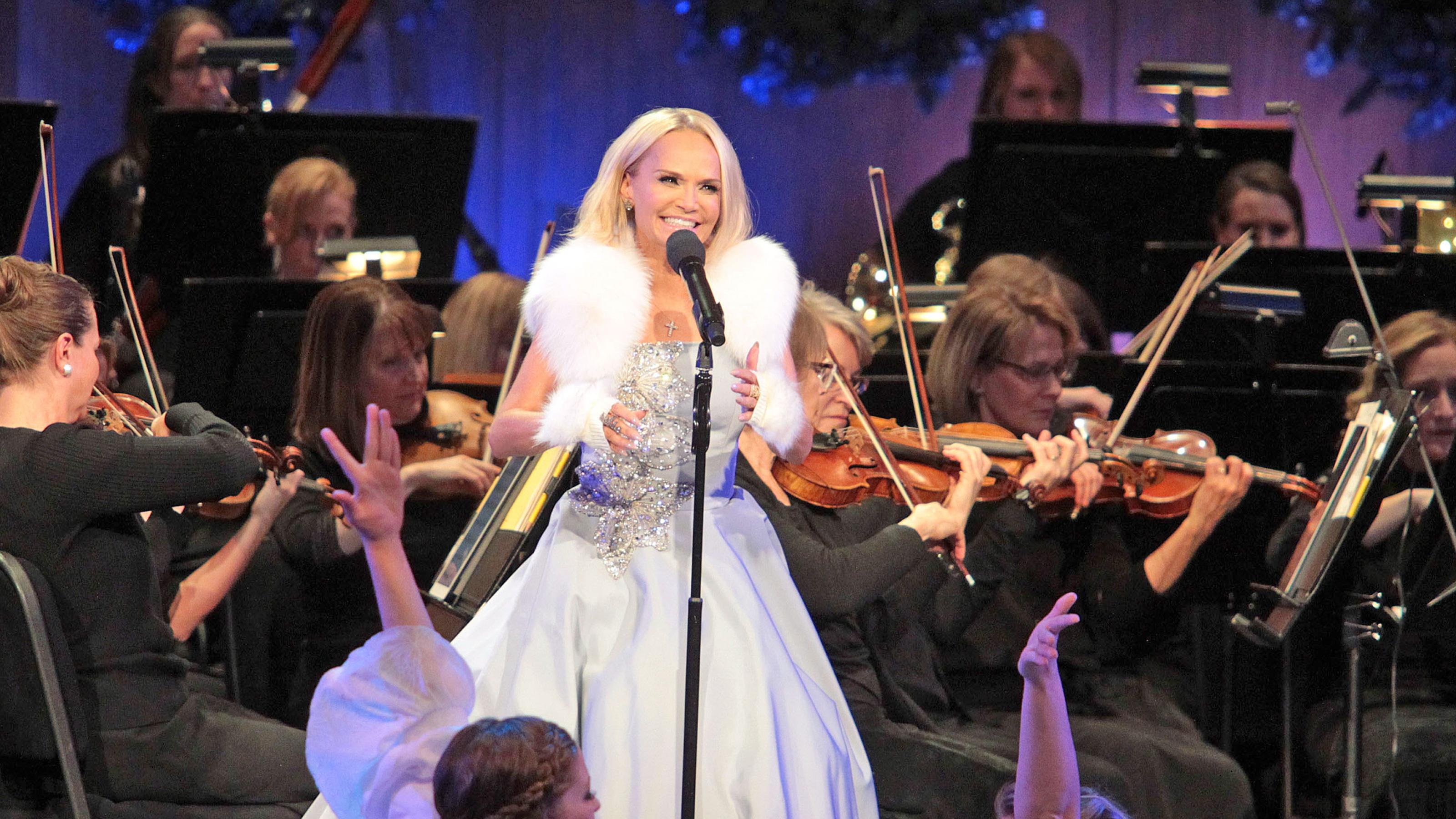 Kristin smiles on stage in front of a microphone with violinists playing behind her.