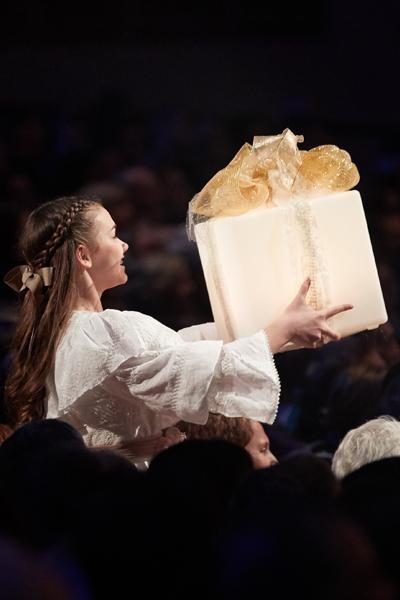 A young woman dressed in white holds up a wrapped, cream-colored present.