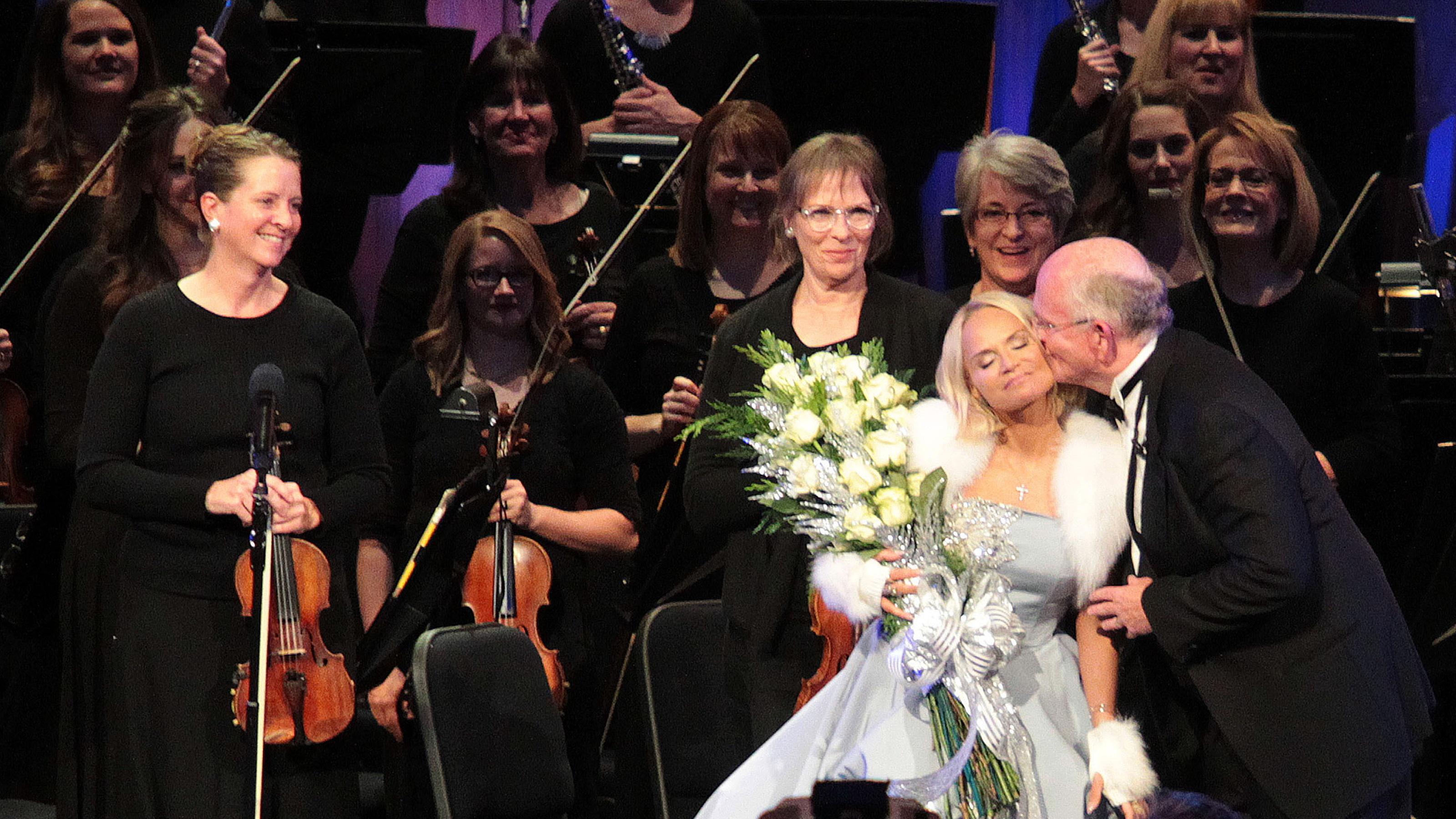 Musicians stand in the background while Mack Wilberg kisses Kristin Chenoweth on the cheek in front of them.