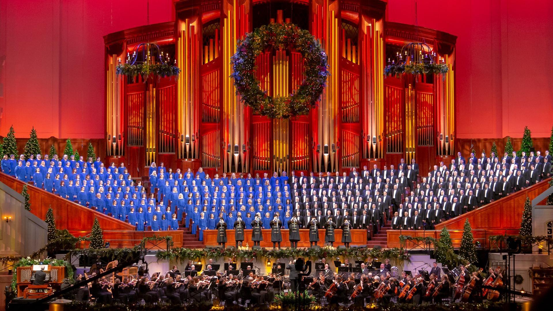 The Tabernacle Choir performs alongside conductor Mack Wilberg and the Orchestra at Temple Square in front of a large organ and festive Christmas decorations.