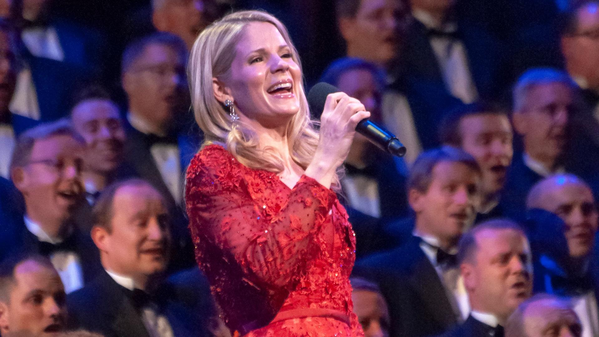 Kelli O'Hara in a red dress sings into a microphone with the choir behind her