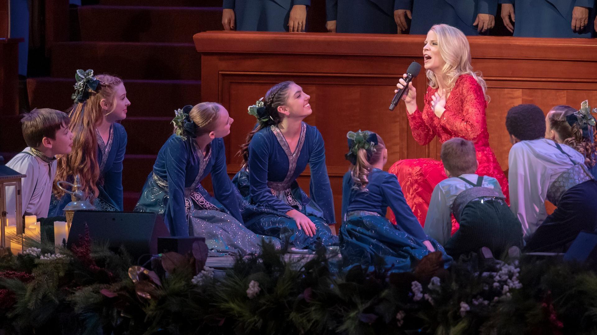 Kristin Chenoweth sings into a microphone amidst the female members of The Tabernacle Choir, who are all dressed in white with single-color scarves.