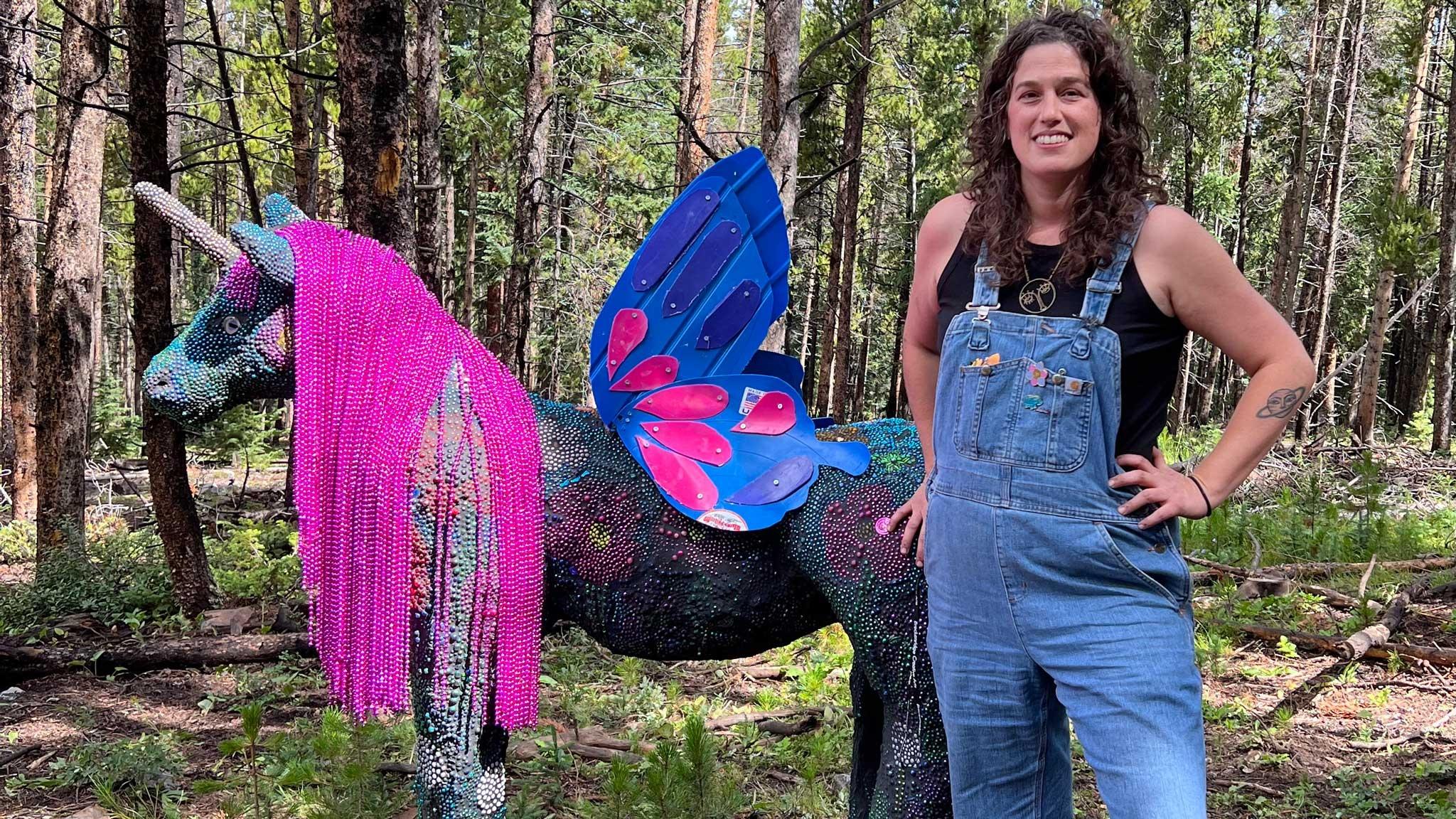 Artist Calder Kamin stands in a forest with her unicorn made completely out of trash, featured in the PLAY episode of Craft in America
