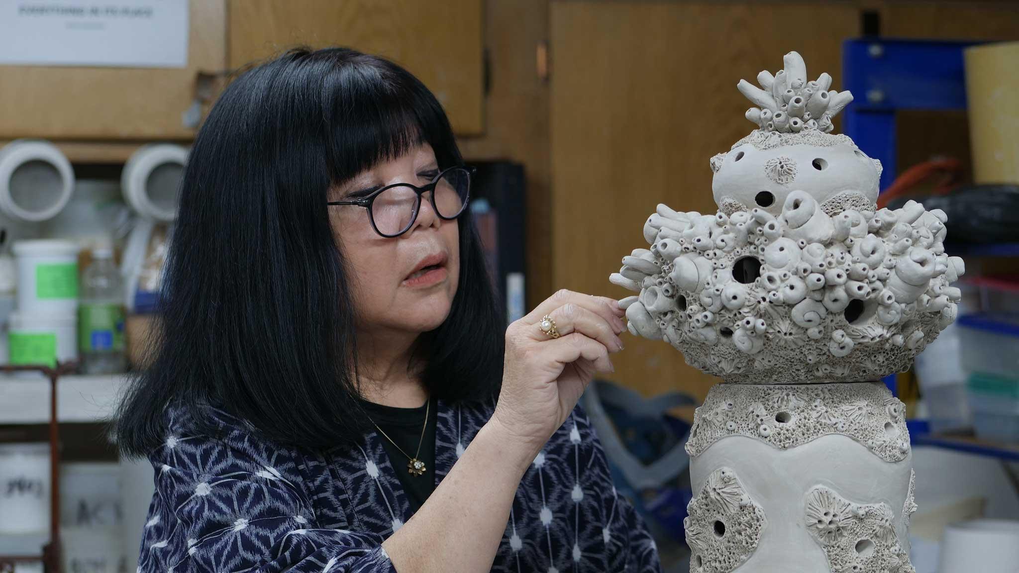 Japanese American woman in blue/white pattern shirt working on coral artworks made out of clay