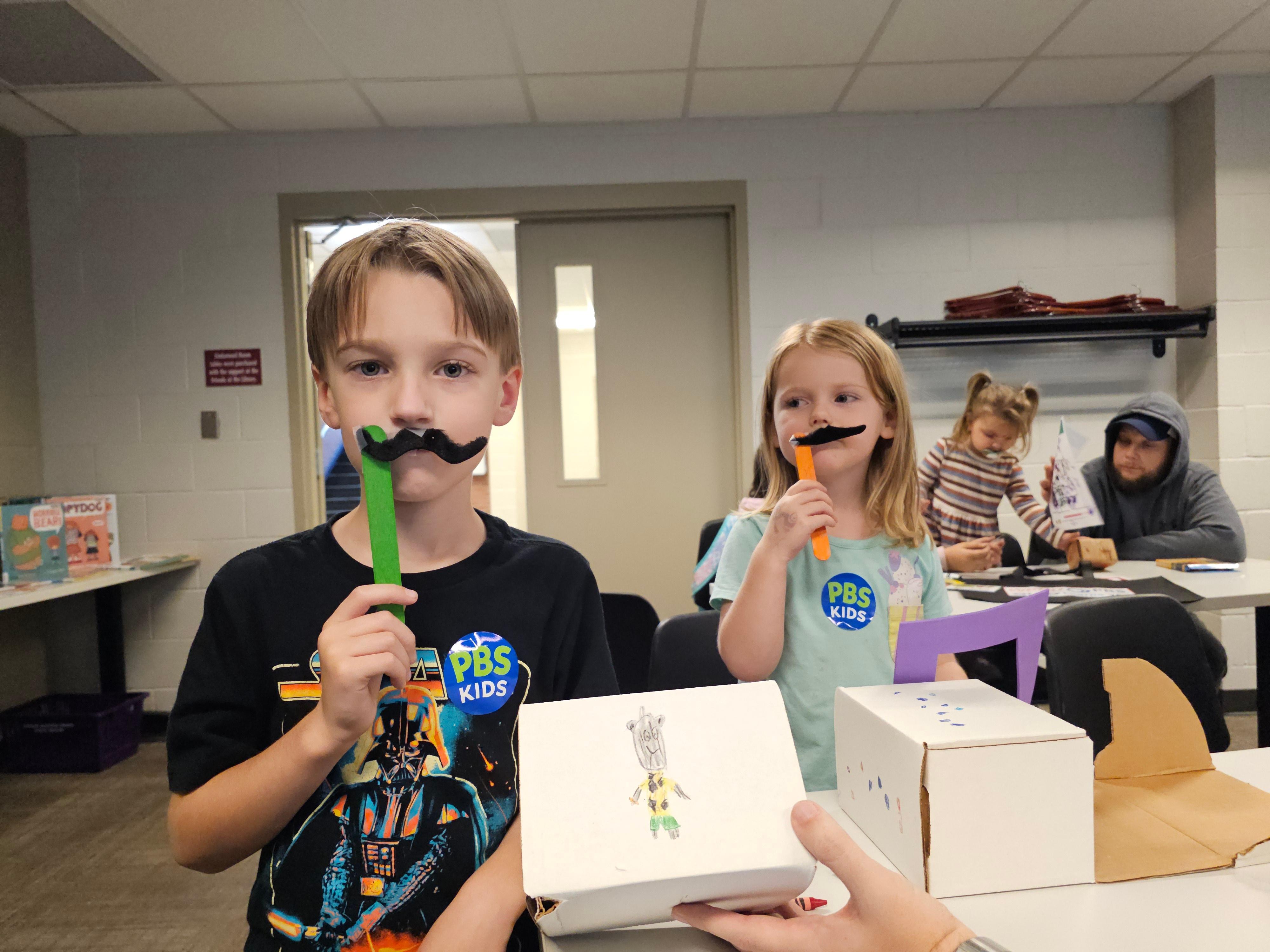 A boy and girl holding fake mustaches up to their faces while showing their collection boxes.