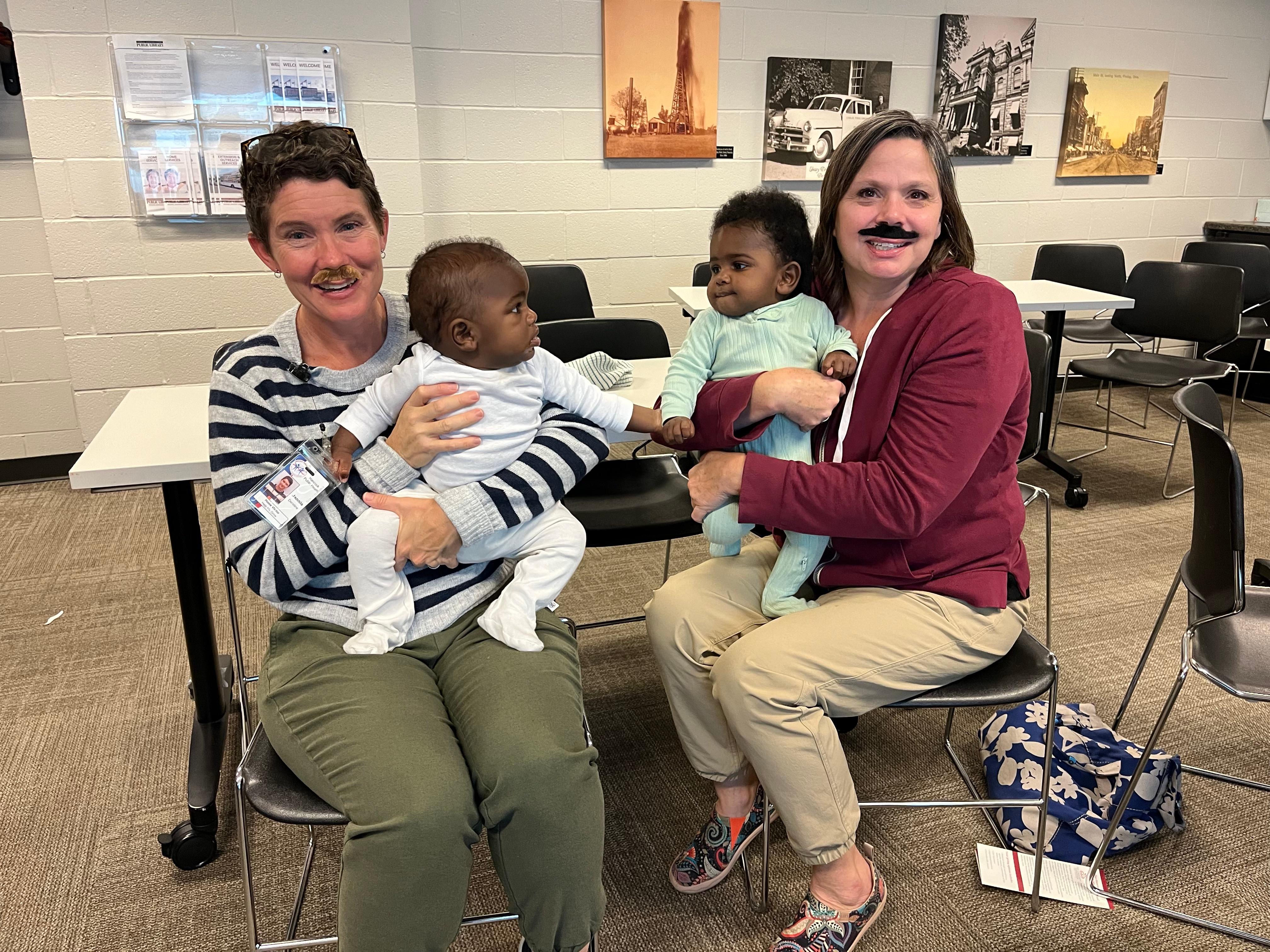 2 women holding babies while wearing mustaches smiling.