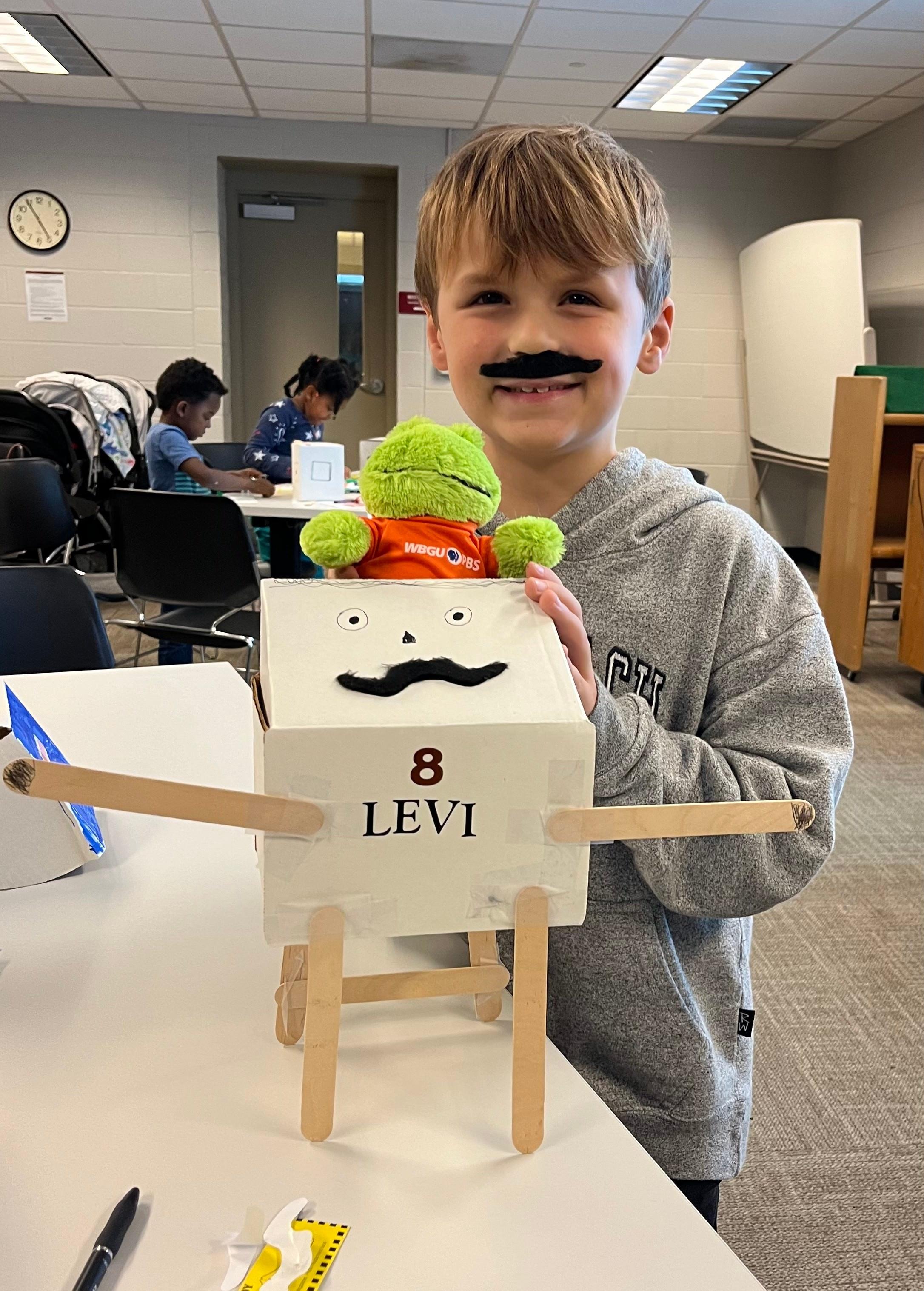 Boy smiling with mustache while holding frog in front of mustache-faced collection box.