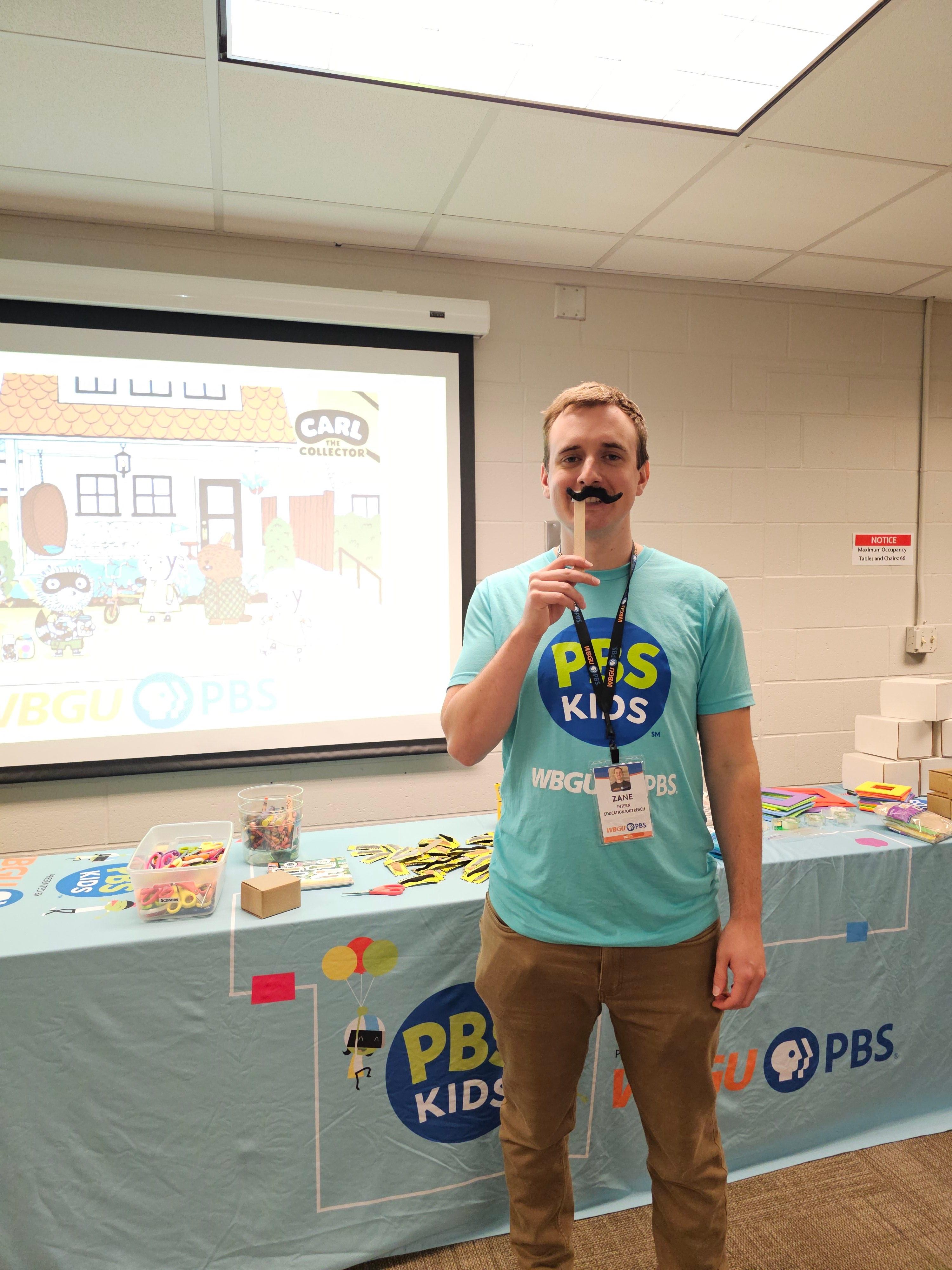 PBS presenter showing off fake mustache craft for the kids.