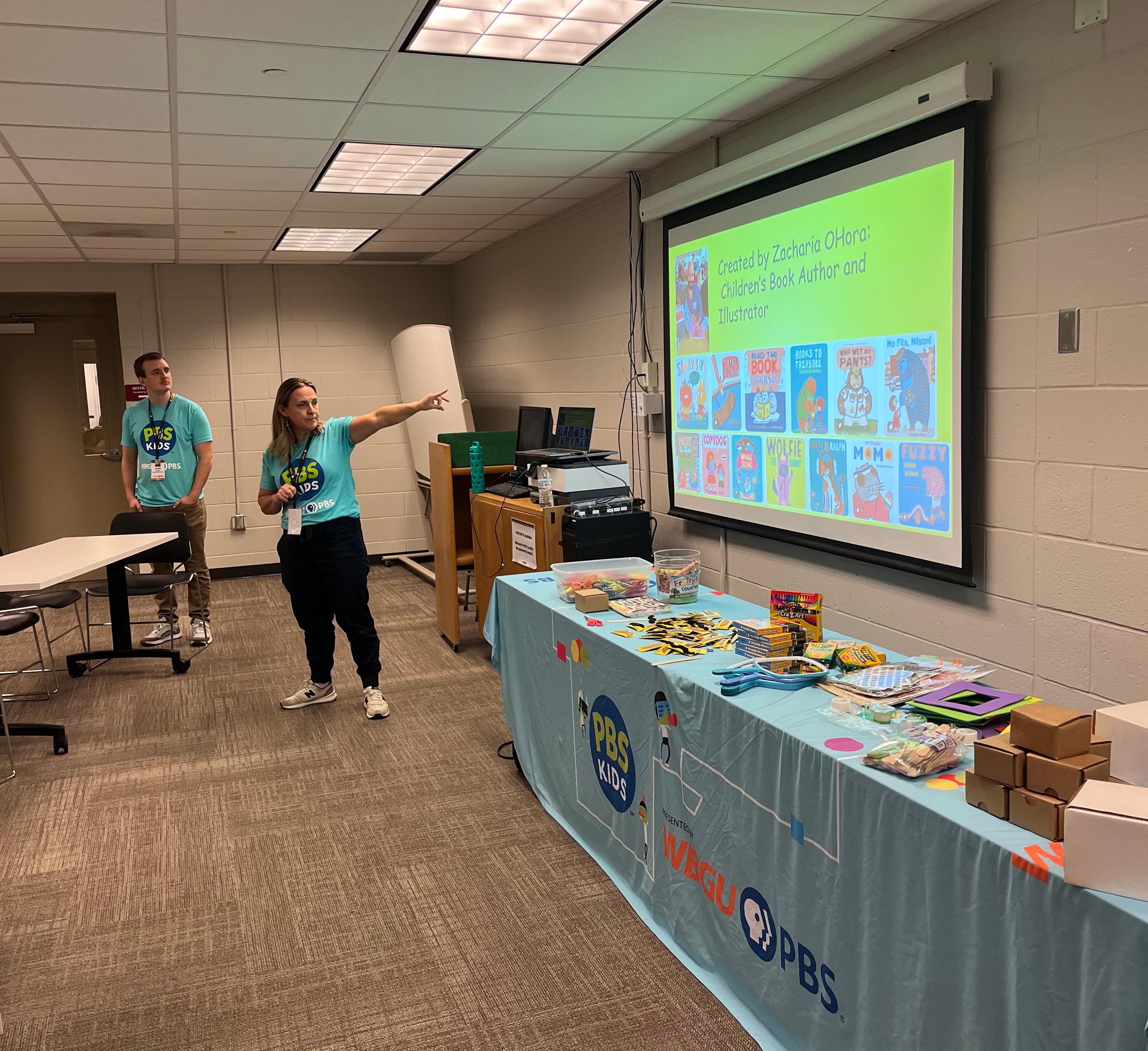 2 PBS KIDS presenters showing Books written by Zacharia O'Hara in front of table of craft supplies.