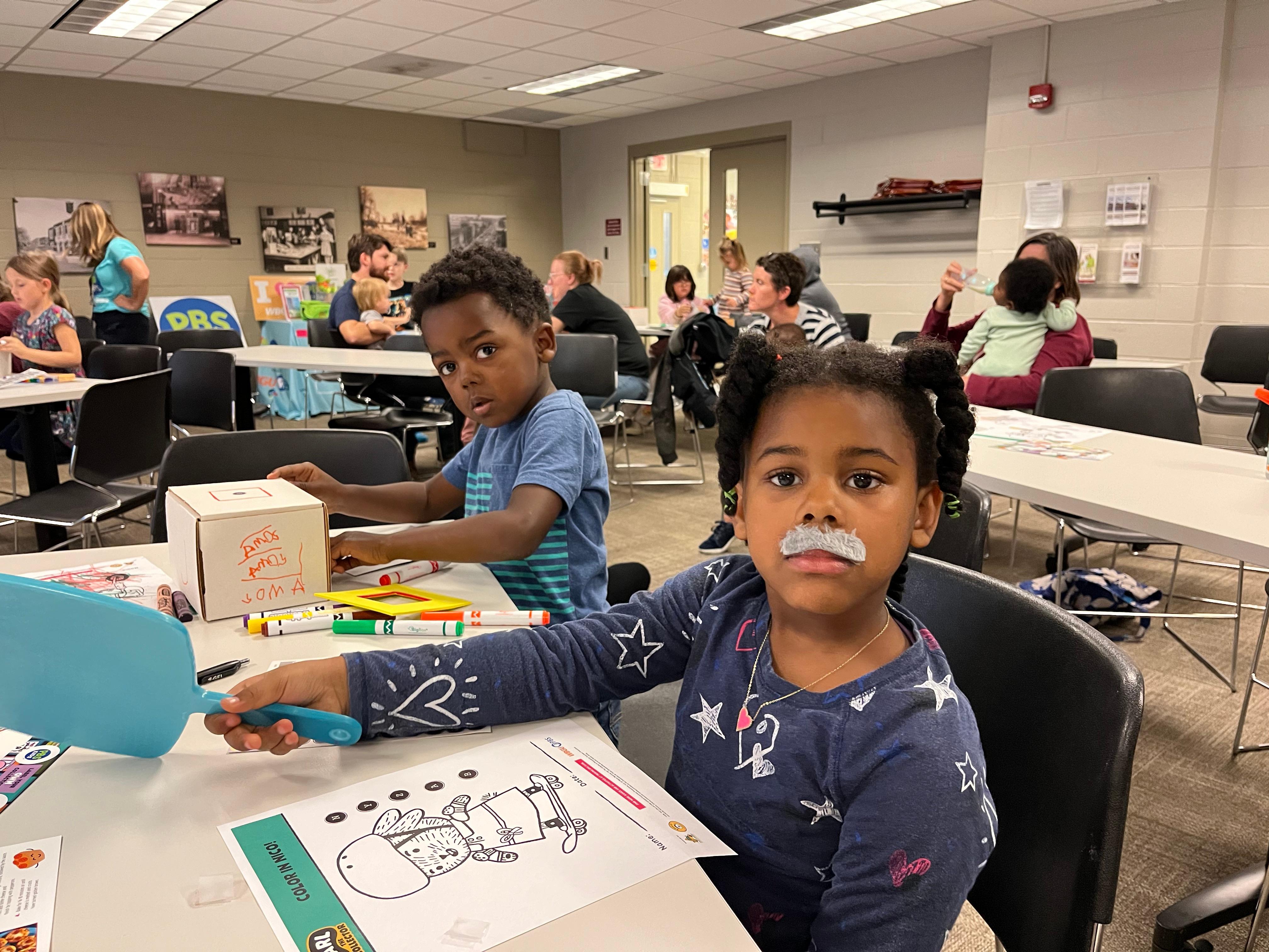 2 kids looking at camera with mustaches on one of their faces while drawing and working on collection box.