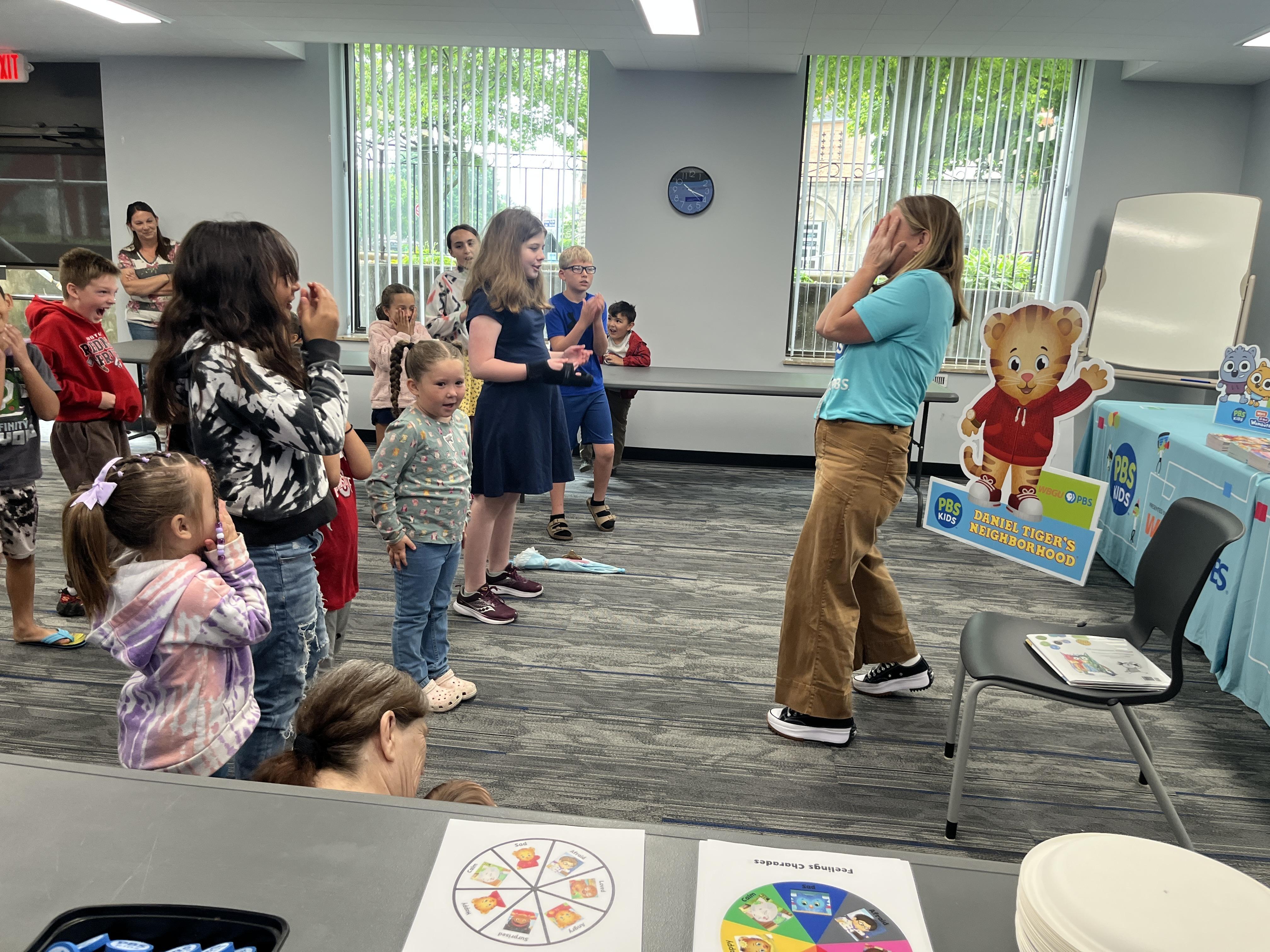 A PBS Kids presenter stands with her hands covering her face in front of a large standing crowd of excited children. They are playing a game where they have to act out an emotion in a creative and visual way with their body language. 