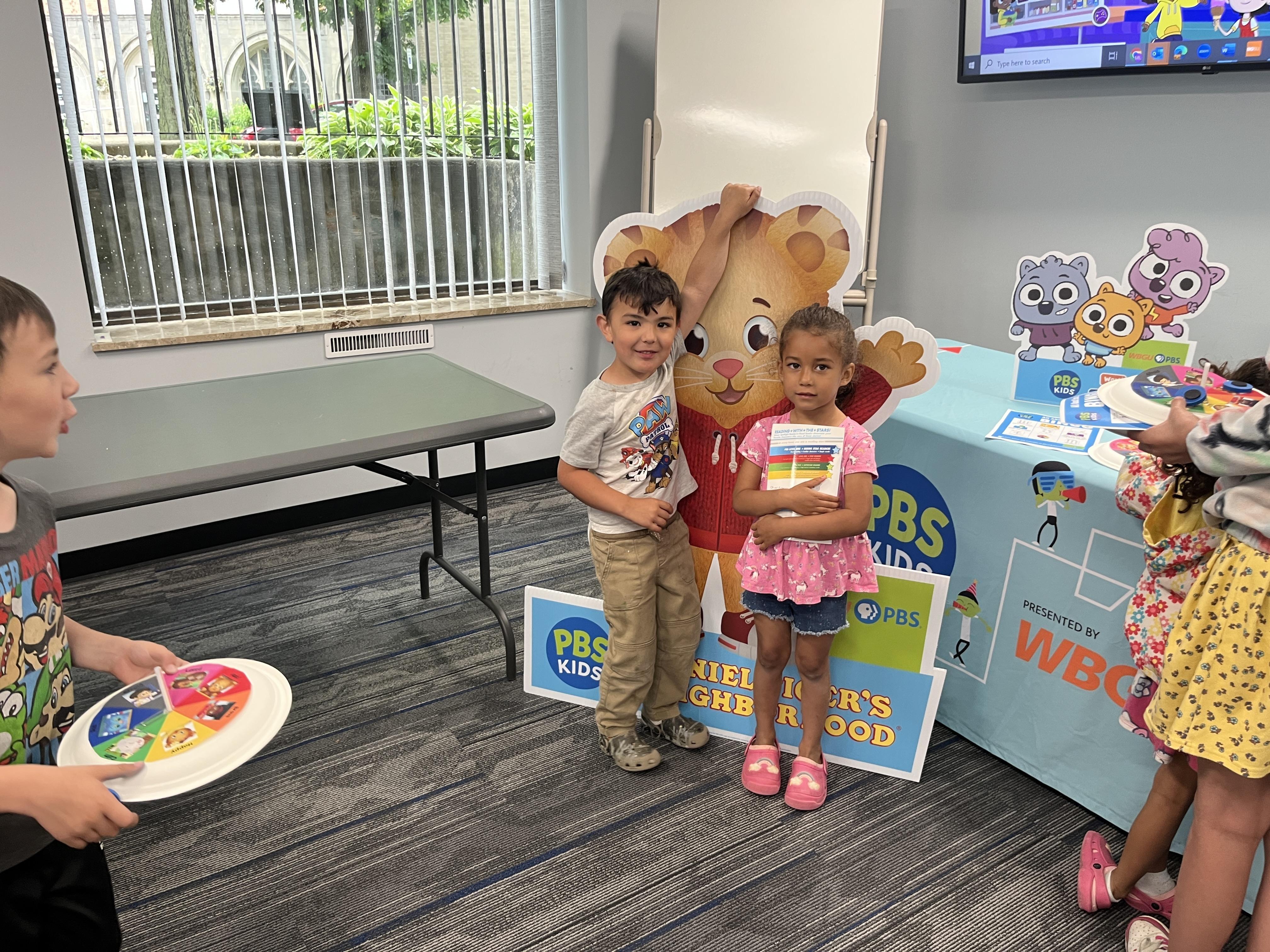 A young boy and girl pose for a picture in front of a "Daniel Tiger's Neighborhood sign." The boy is holding onto the top of the sign and the girl is clutching a book with both hands. In the left foreground another boy is holding an emotions spinner craft.