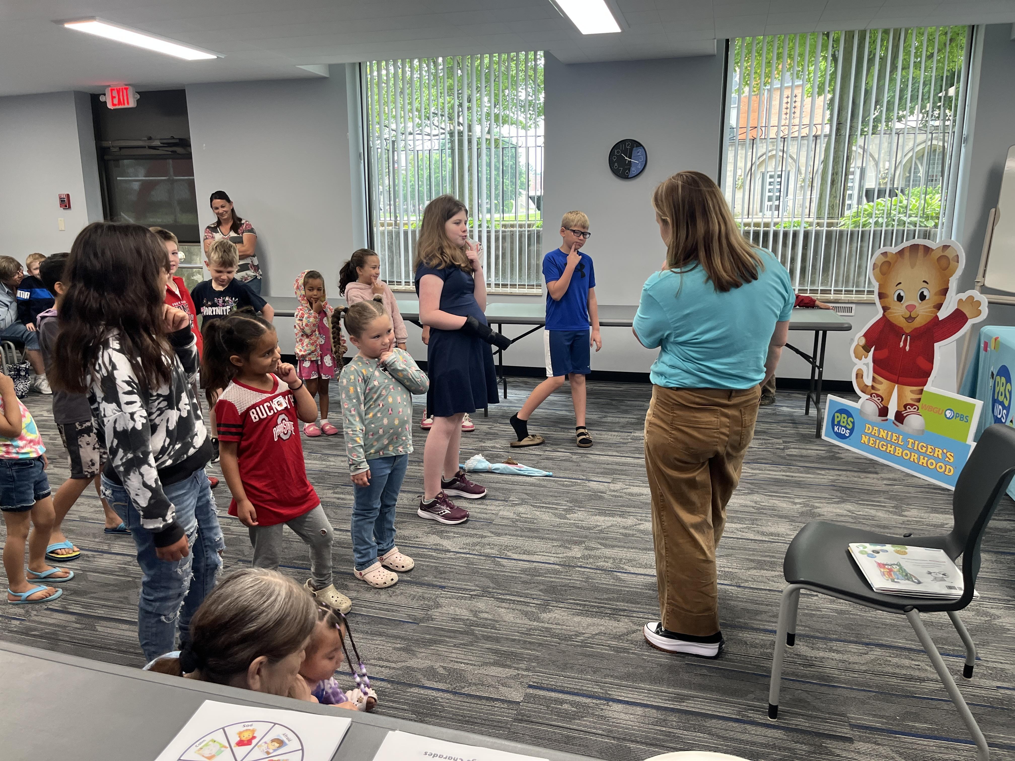 A PBS presenter playing an emotions and body language game with a group of excited children. They are acting out what it is like to be curious with a finger on their chin and inquisitive look on their faces.