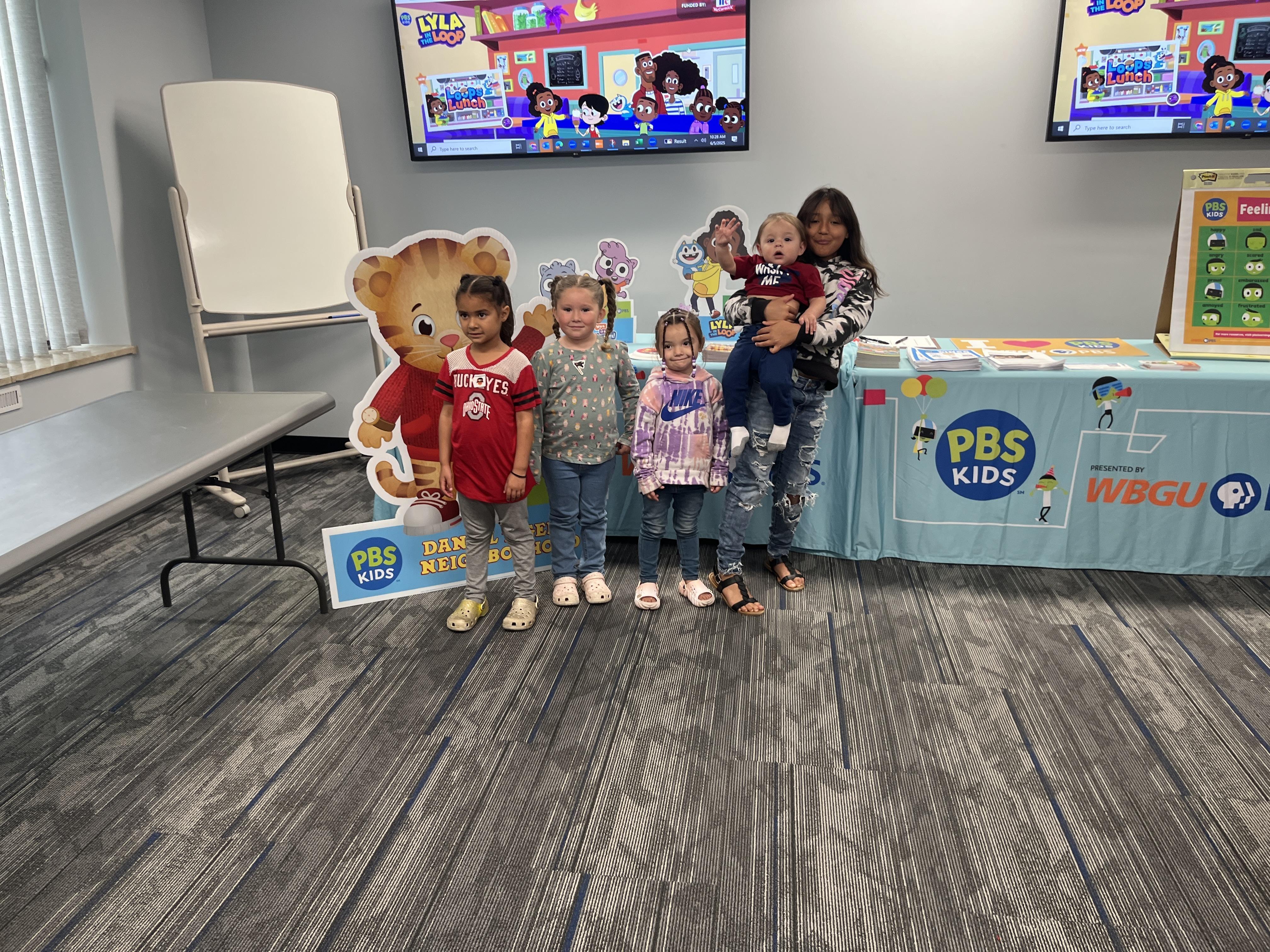 A group of five children all standing for a picture in front of a "Daniel Tiger's Neighborhood display and PBS Kids and WBGU-PBS table full of character displays, and emotions poster and packets. Three young girls are standing next to each other and one larger girl is holding onto a very young boy who is holding his hand up. A "Lyla in the Loop" website is displayed on the TV's behind them.