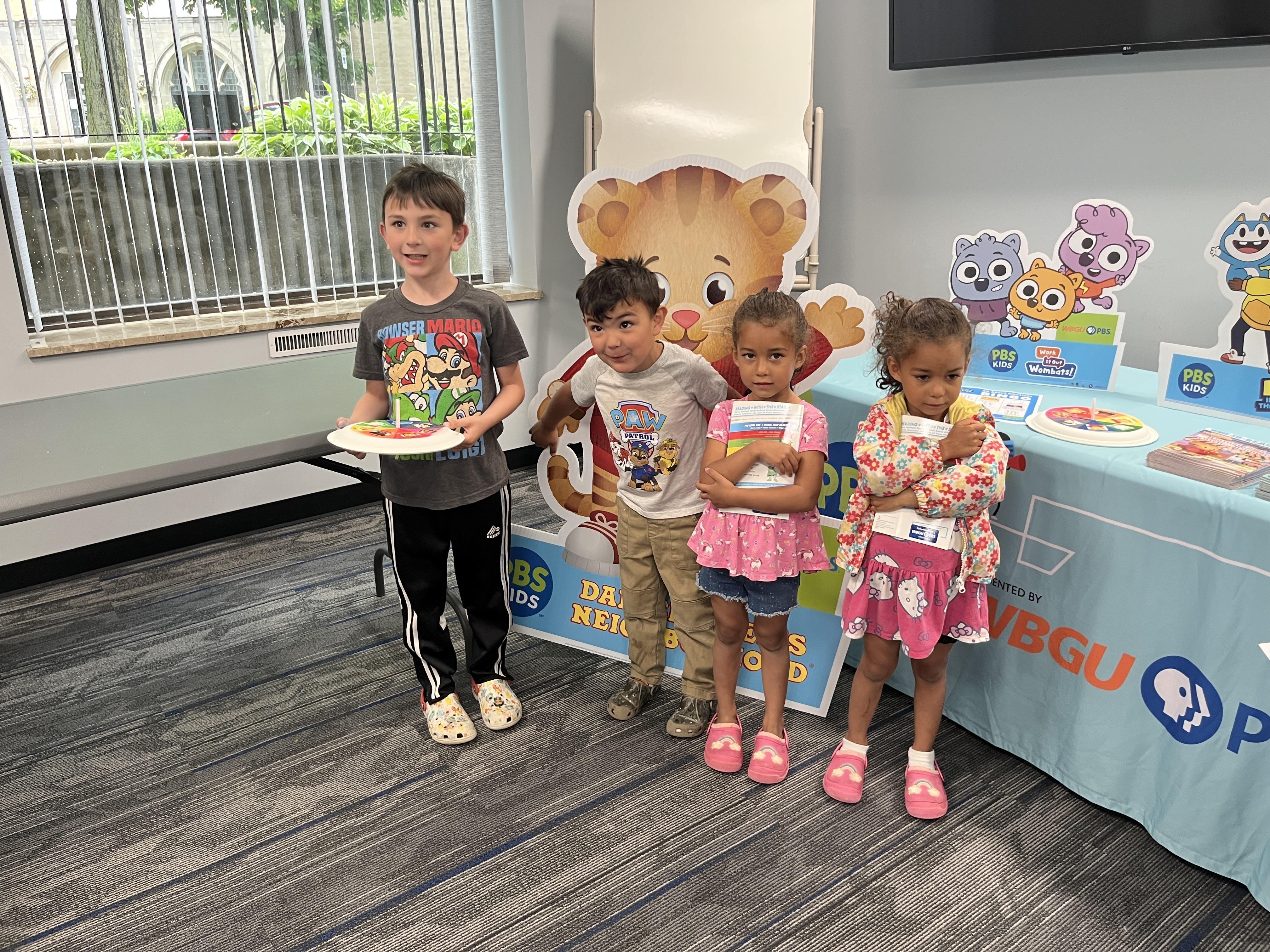Four children standing in front of a "Daniel Tiger's Neighborhood" display.  One boy is holding a spinner and smiling the other is holding onto the display with both hands behind himself. Both girls are standing next to each other to the right and are clutching a book.