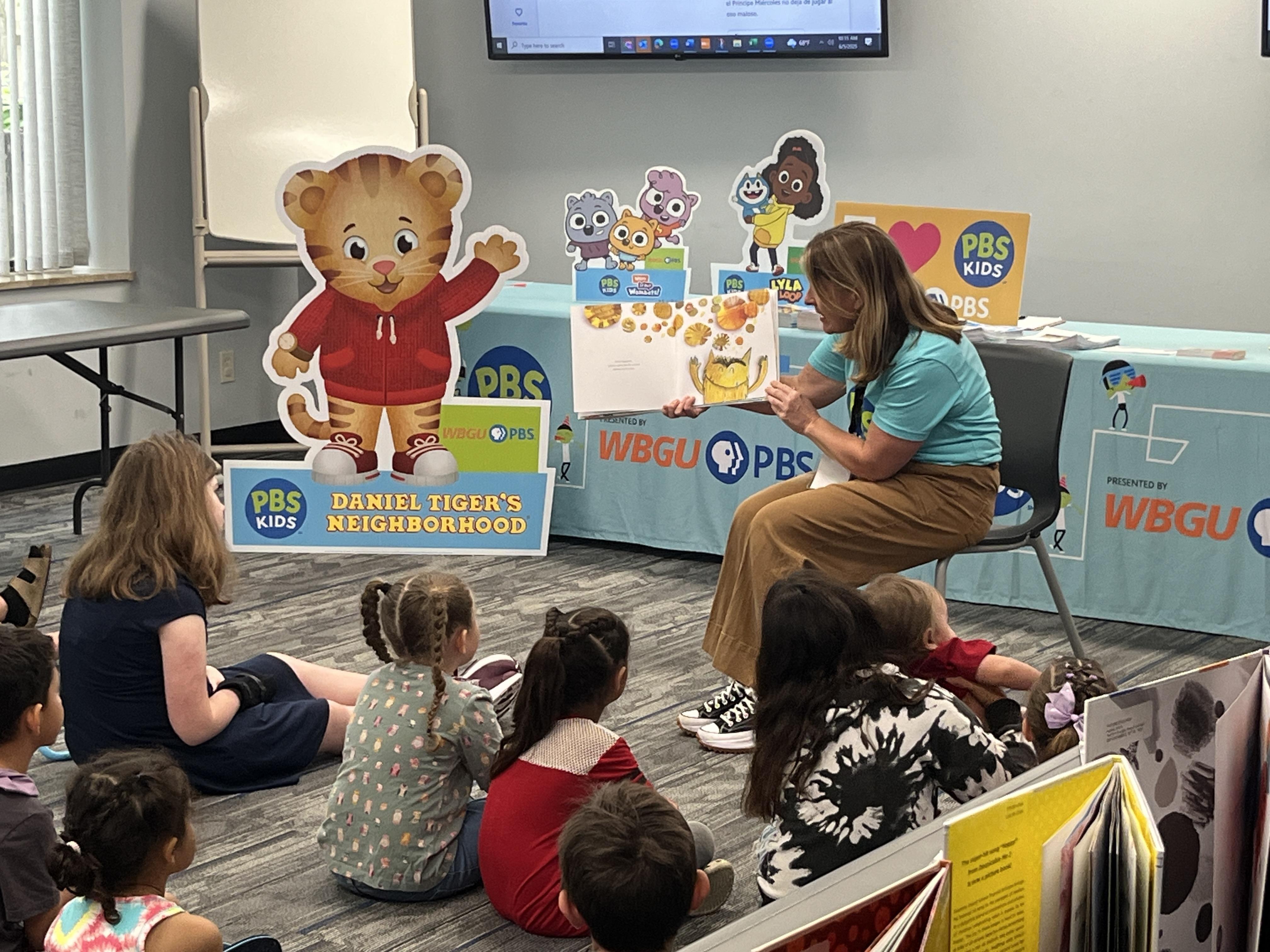 A zoomed in shot of a PBS Kids presenter reading a book about emotions to a group of young children sitting on the floor. A "Daniel Tiger's Neighborhood" sign is along with other promotional displays are in the background along with a WBGU-PBS table.