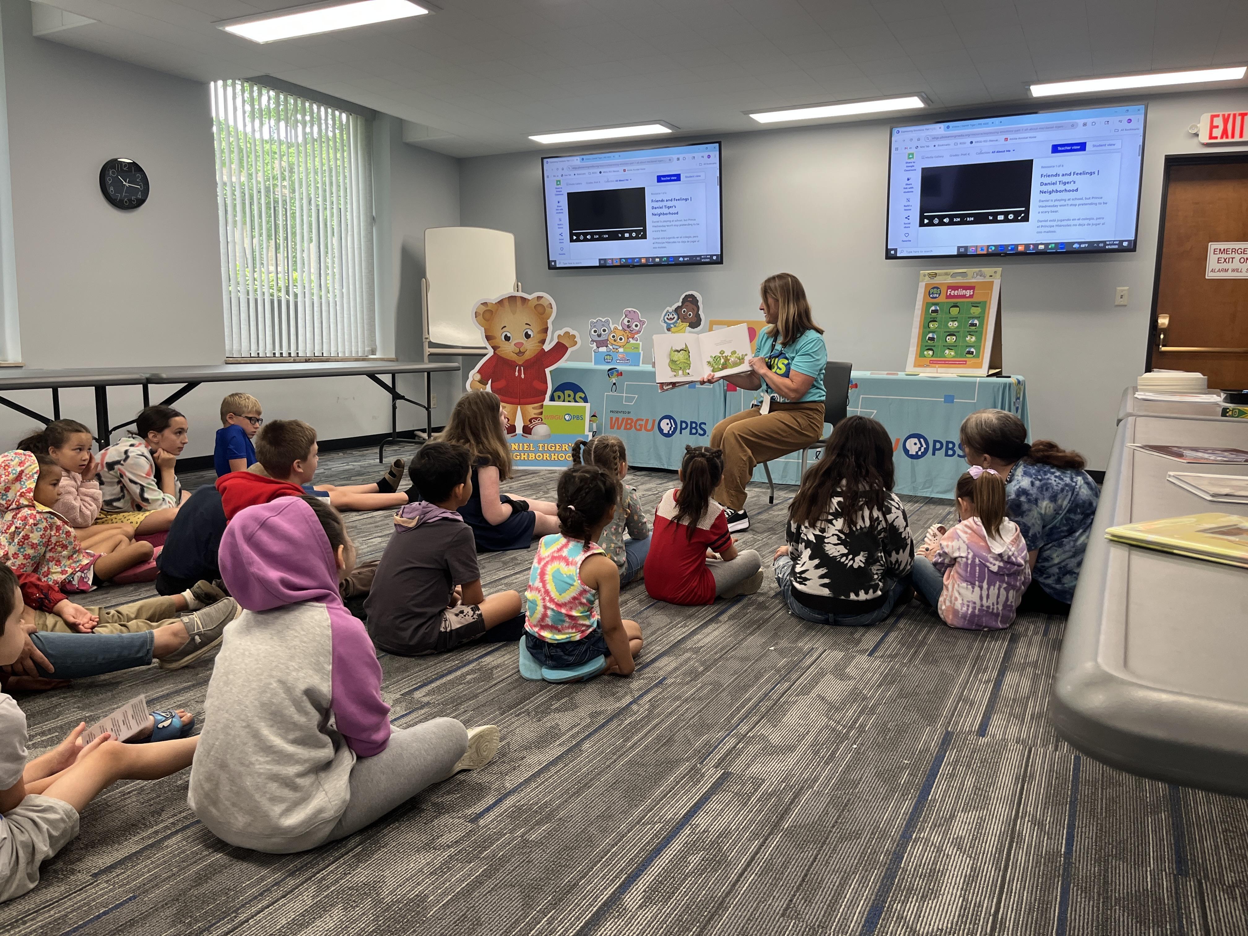 A wide shot of a PBS Kids presenter reading a book about emotions to a group of young children sitting on the floor. A "Daniel Tiger's Neighborhood" sign is along with other promotional displays are in the background along with a WBGU-PBS table.