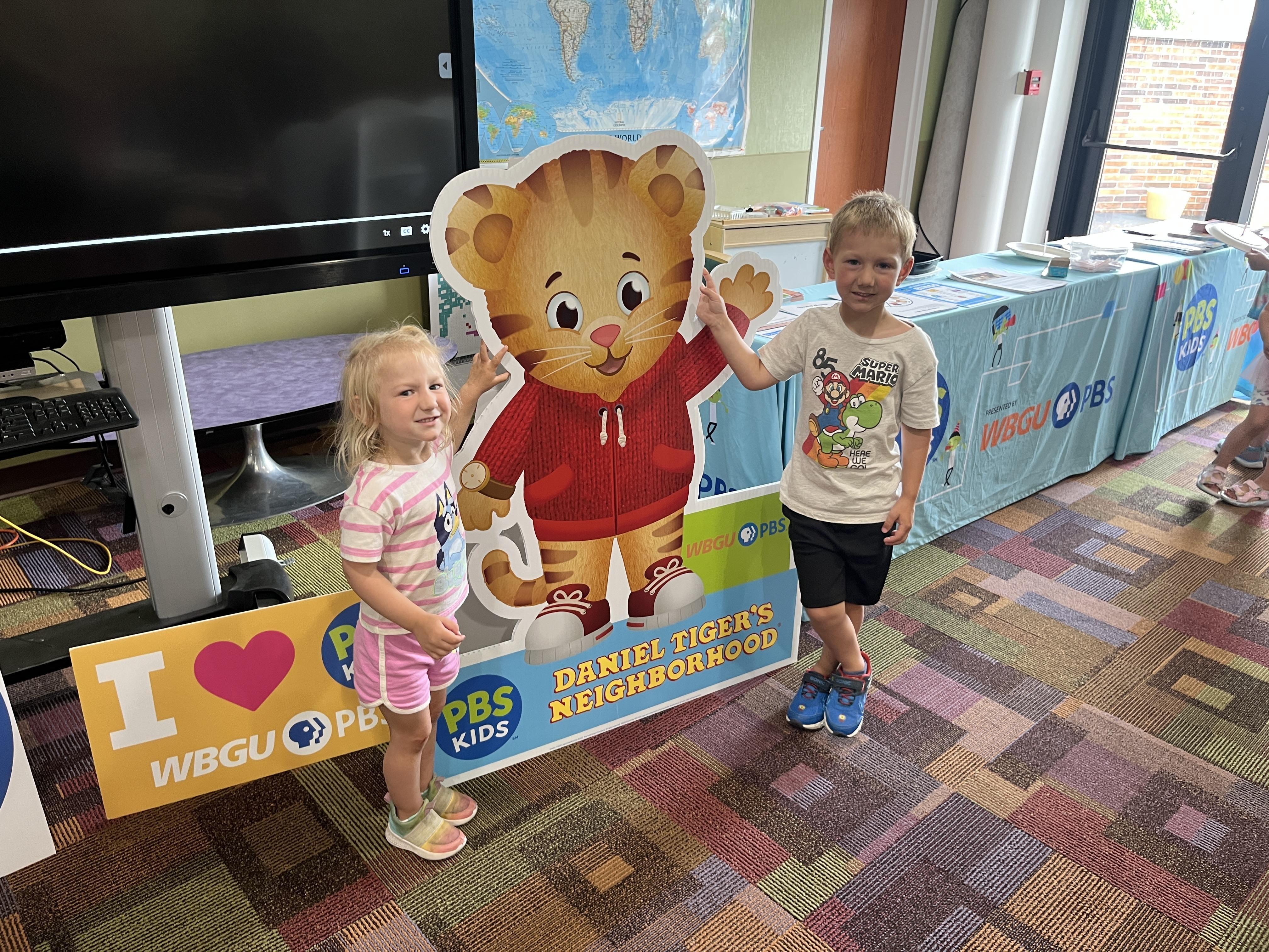 A young girl in a pink and white striped shirt and pink shorts, and a young boy in a white Super Mario t-shirt and black shorts, stand on either side of a large cardboard cutout of Daniel Tiger. They are all standing on a colorful patterned rug. In the background, there's a large screen, a world map on the wall, and a table with PBS Kids branding.