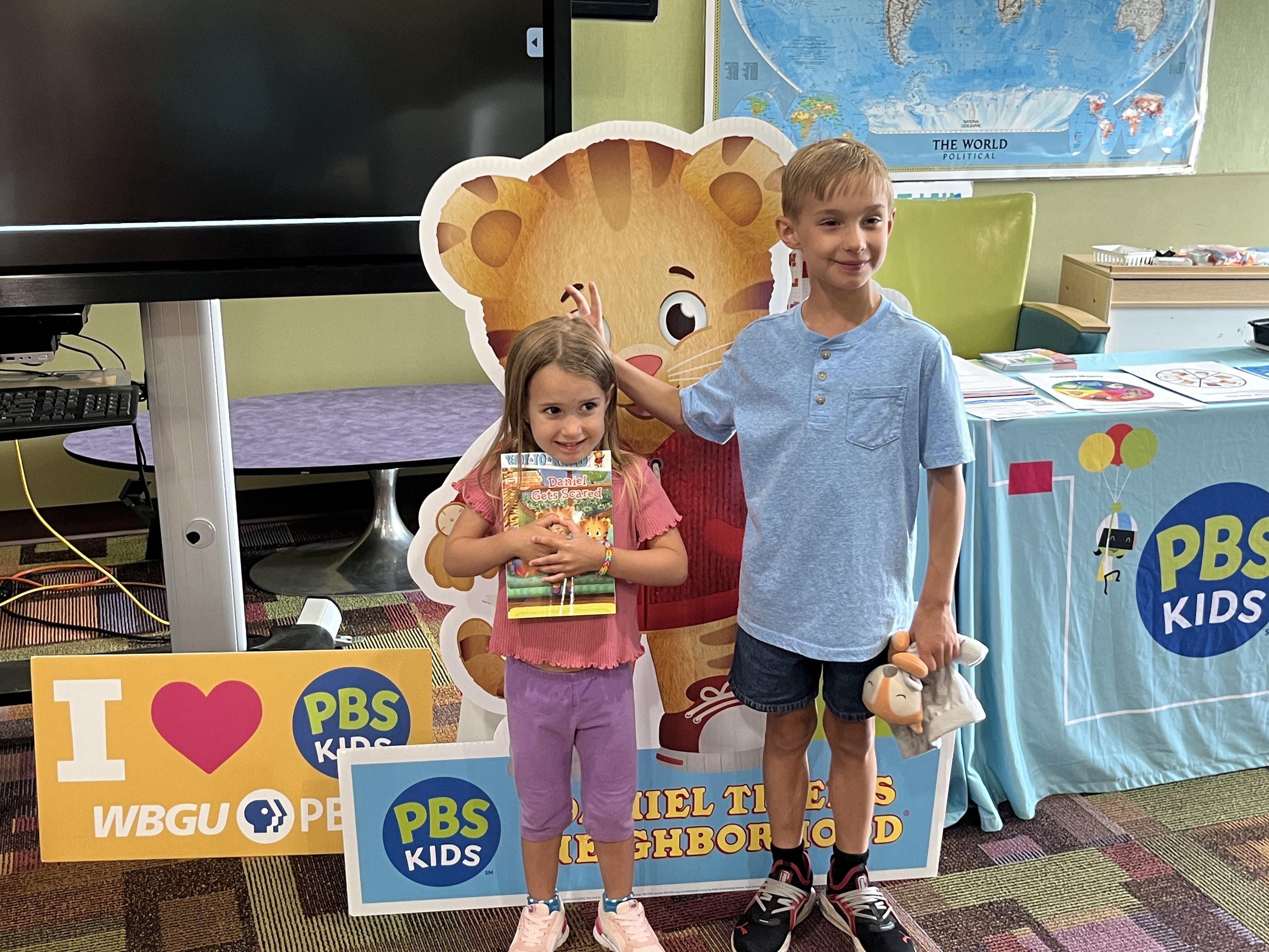 A young girl in a pink shirt and purple pants smiles and holds a book, while a taller boy in a blue shirt and jean shorts stands next to her, playfully holding his hand above her head, as if giving her "bunny ears." They are standing in front of a large Daniel Tiger cutout. A world map hangs on the wall in the background, and a PBS Kids branded table is visible to the right.