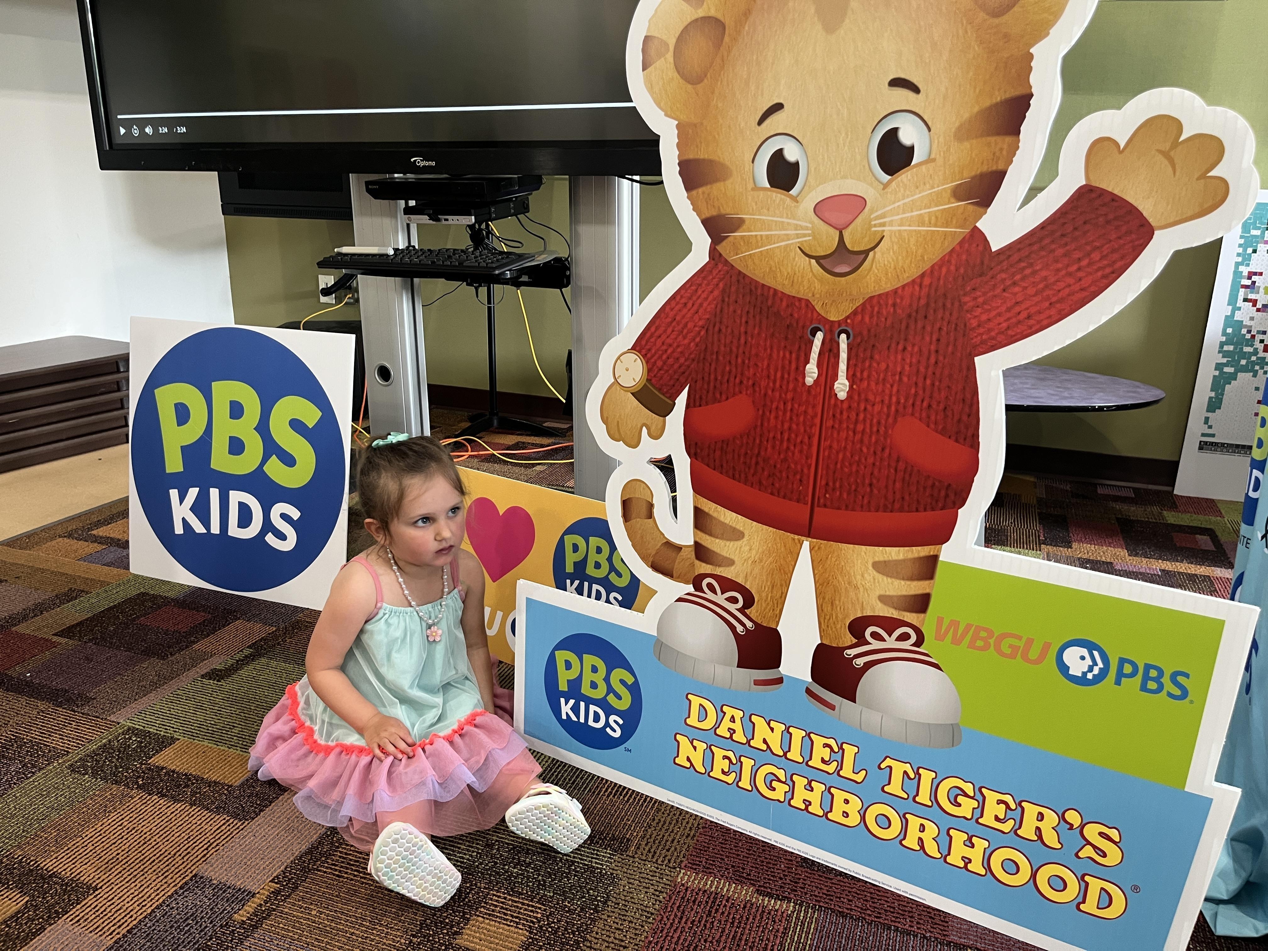A young girl with light brown hair sits on a colorful patterned carpet in front of a large cardboard cutout of Daniel Tiger from "Daniel Tiger's Neighborhood." She is wearing a light blue top and a pink tutu-style skirt, with white shoes. Several PBS Kids logos and a "Daniel Tiger's Neighborhood" sign are also visible around her. In the background, there is a large flat-screen monitor or TV on a stand.