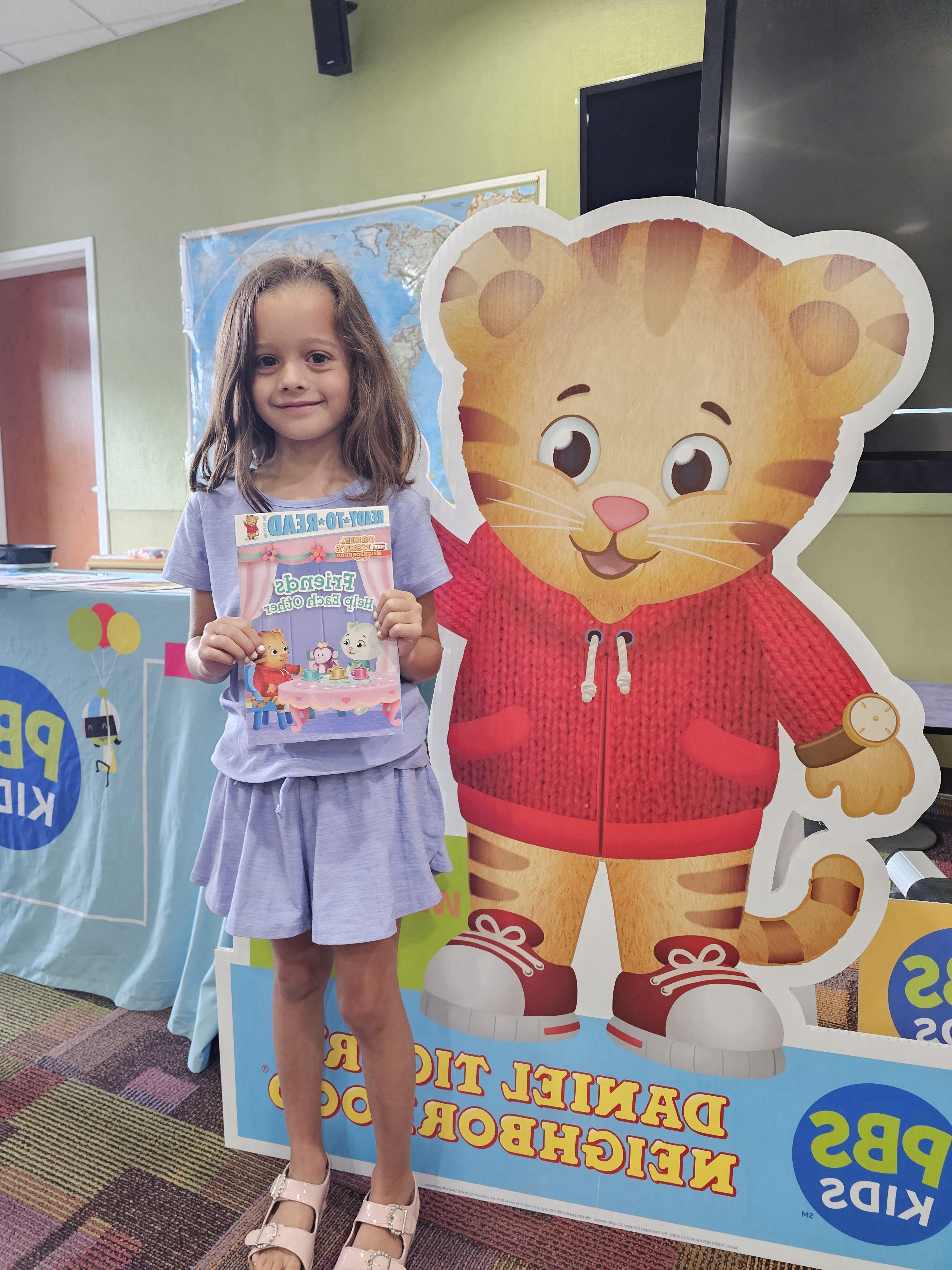 A young girl with long brown hair, wearing a light purple dress and pink sandals, smiles as she holds up a book titled "Friends Help Each Other" featuring Daniel Tiger characters. She is standing next to a large cardboard cutout of Daniel Tiger. In the background, there's a world map on the wall and a table with PBS Kids branding.