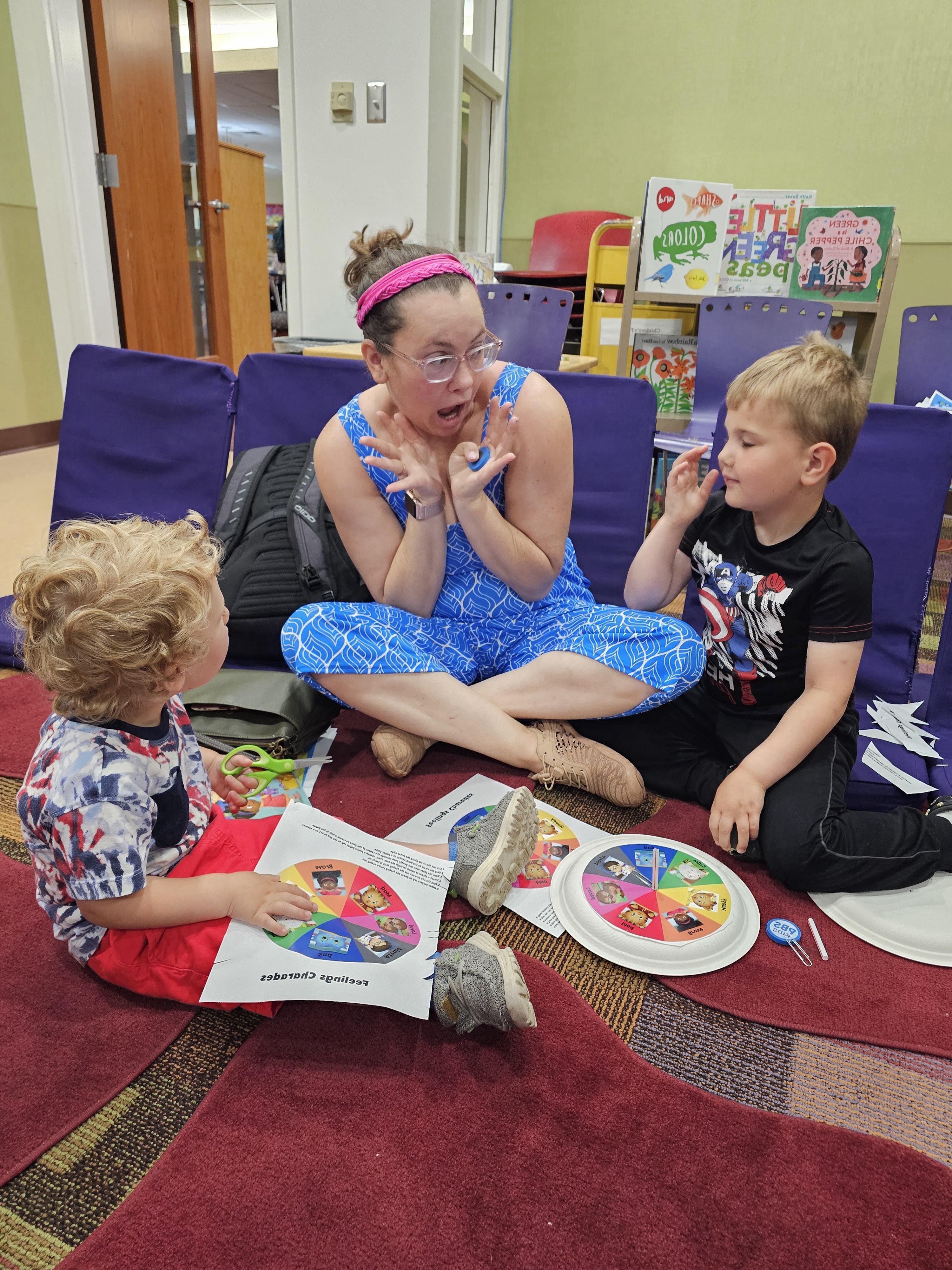  An adult woman with a pink headband and a blue patterned dress sits cross-legged on a dark red rug, looking animated with an open mouth and hands raised, seemingly acting out an emotion. Two young boys are seated on the rug with her. The boy on the left, wearing a black t-shirt and dark pants, is looking at the woman. The boy on the right, with curly blonde hair and a patterned t-shirt and red shorts, is looking at a paper labeled "Feelings Charades." A spinner board with different emotions is also on the rug between them. In the background, there are purple chairs and bookshelves filled with children's books.