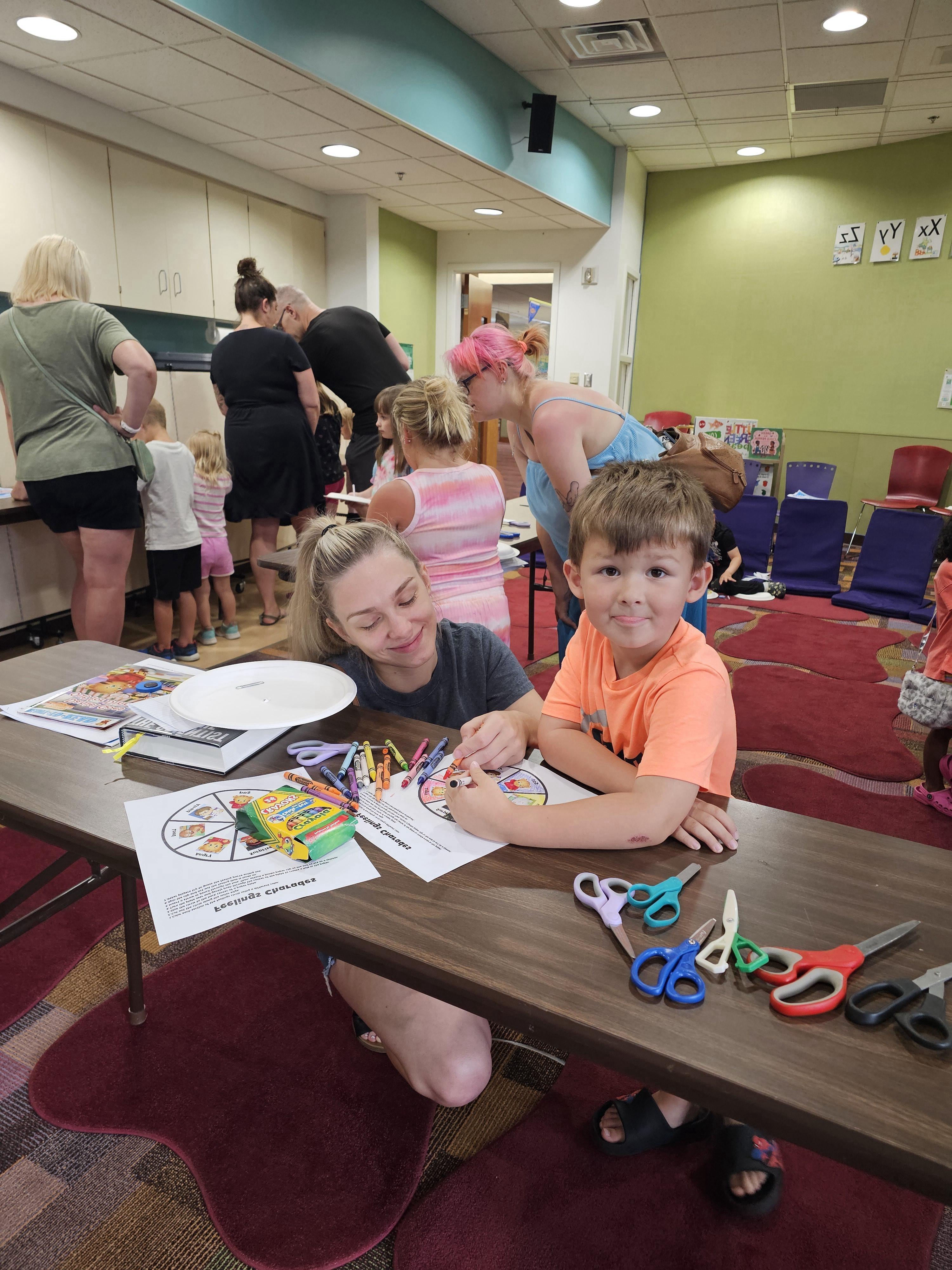 An adult woman with blonde hair and a young boy in an orange t-shirt are seated at a wooden table, engaged in a coloring activity using papers that appear to be part of "Feelings Charades." Numerous pairs of colorful scissors are scattered on the table in front of them. In the background, other children and adults are visible around other tables or standing.