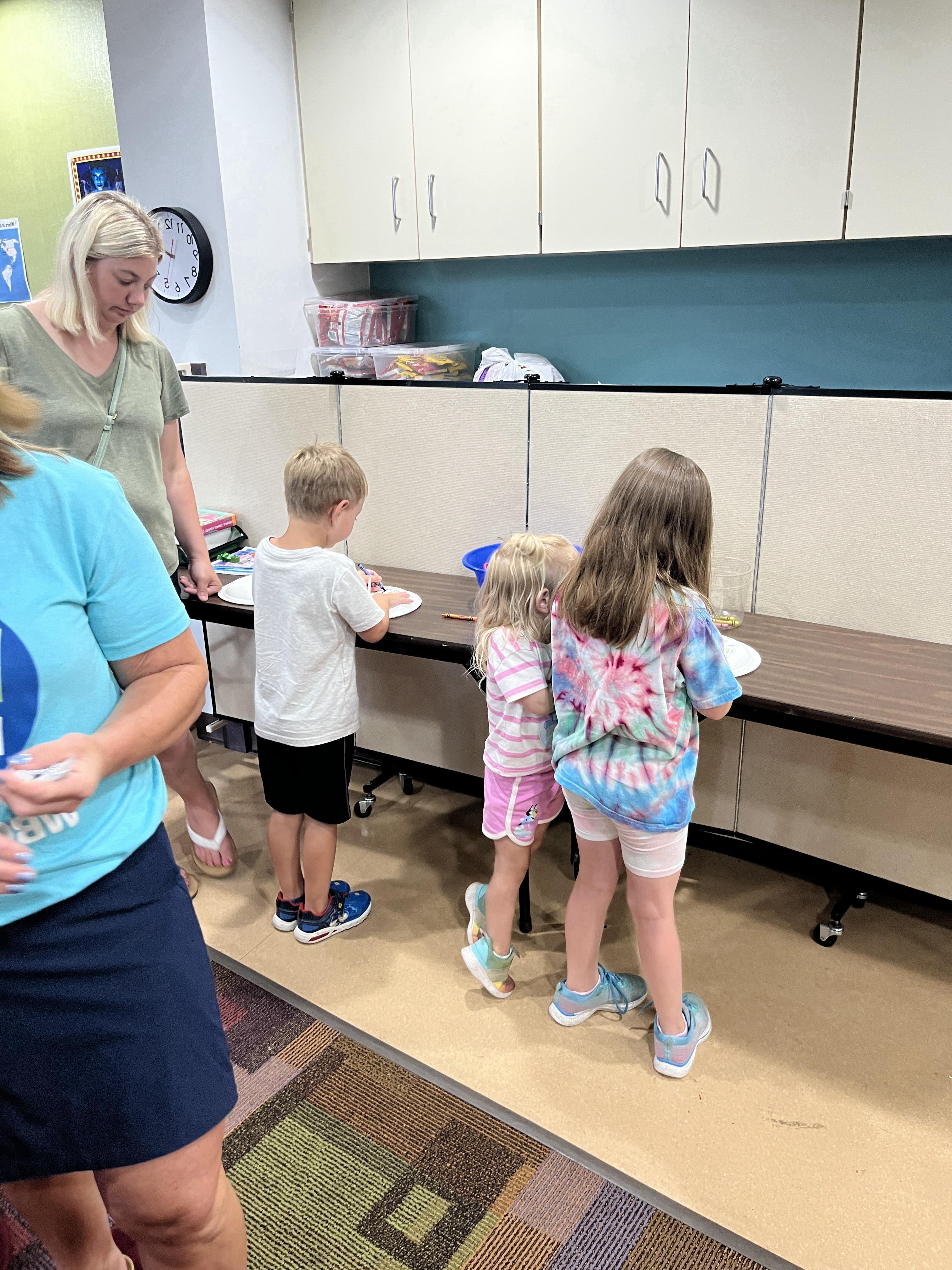 "Three young children, two girls and a boy, are engaged in an activity at a long table, possibly drawing or crafting, with two adult women supervising. The setting appears to be an indoor educational or activity space, featuring white cabinets and a patterned rug."