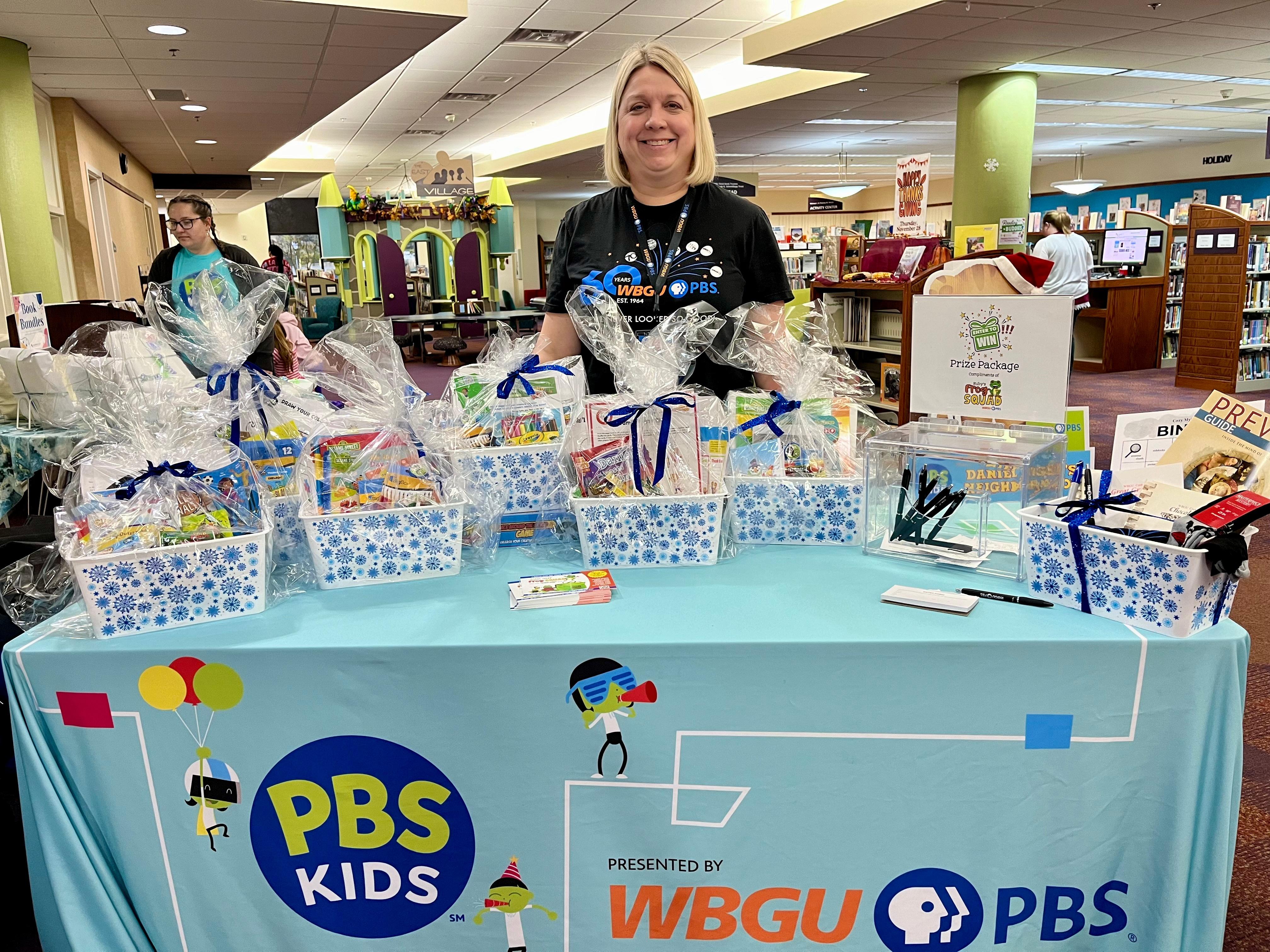 WBGU employee standing in front of gift basket raffle prizes on a table displaying a prominent PBS KIDS and WBGU PBS logo.