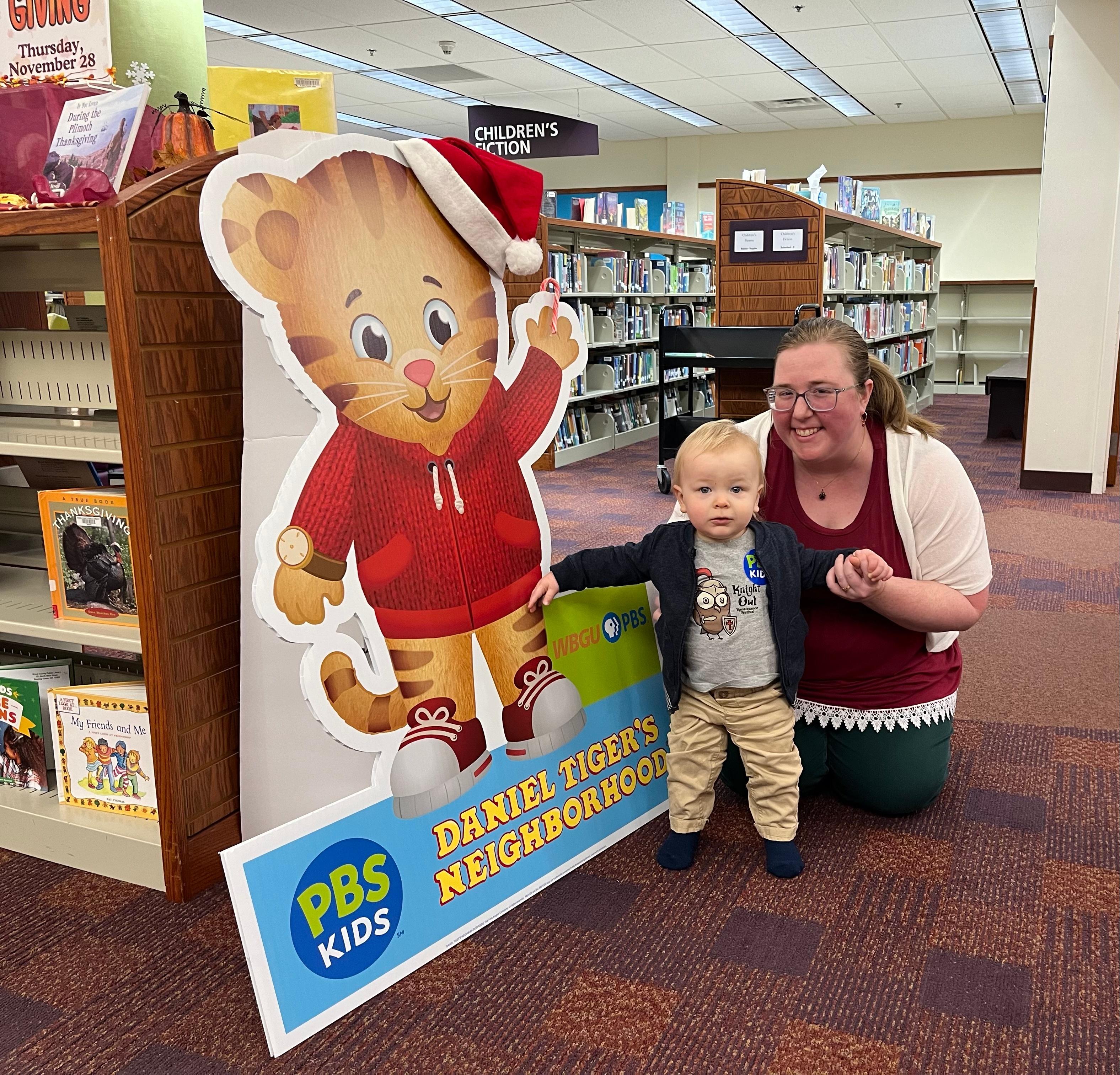 A guardian and young child posing next to a Daniel Tiger's Neighborhood display with PBS KIDS logo in the library.