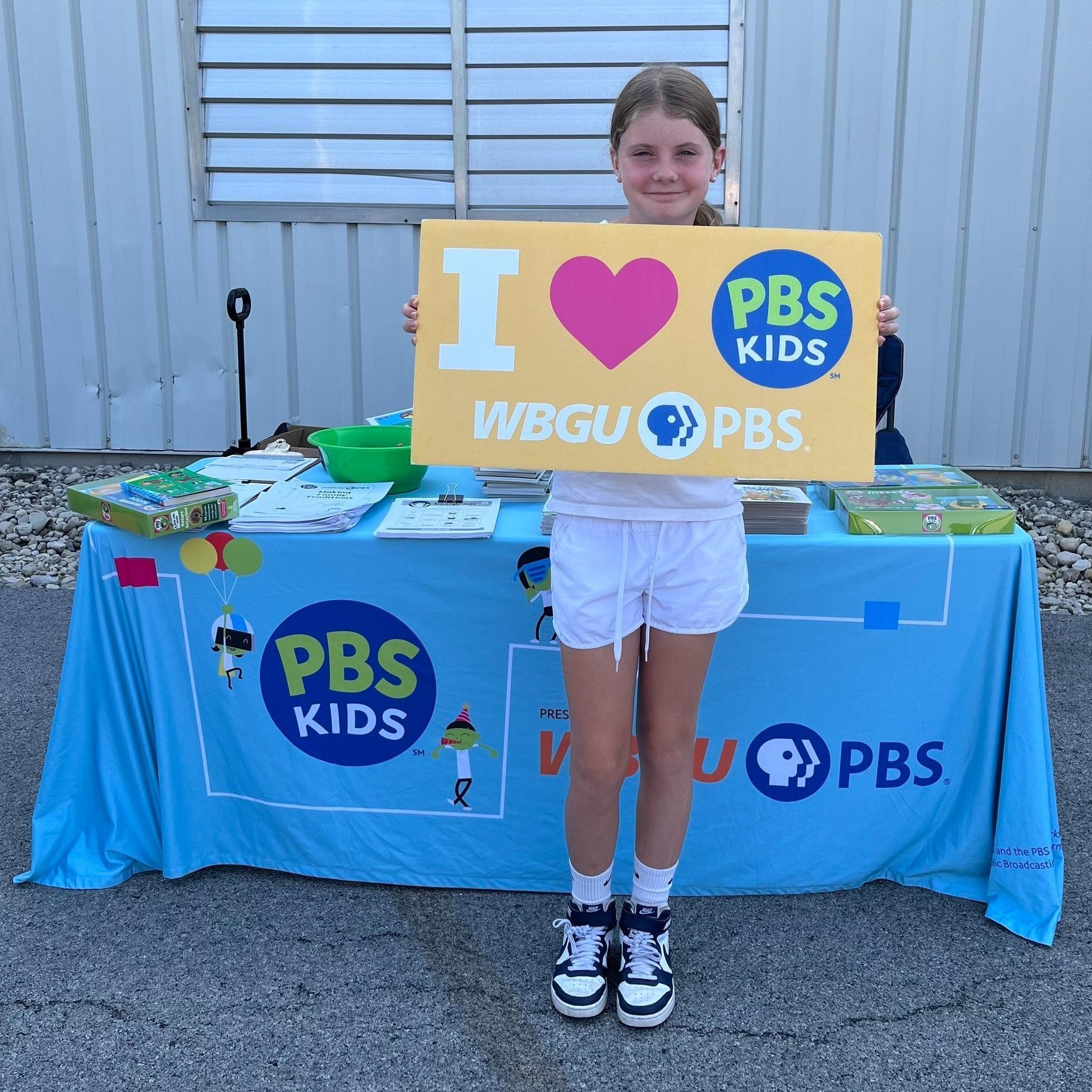 A smiling school aged girl holds a yellow I Love WBGU-PBS sign in front of WBGU's light blue display table in the parking lot of the Findlay YMCA East Branch.