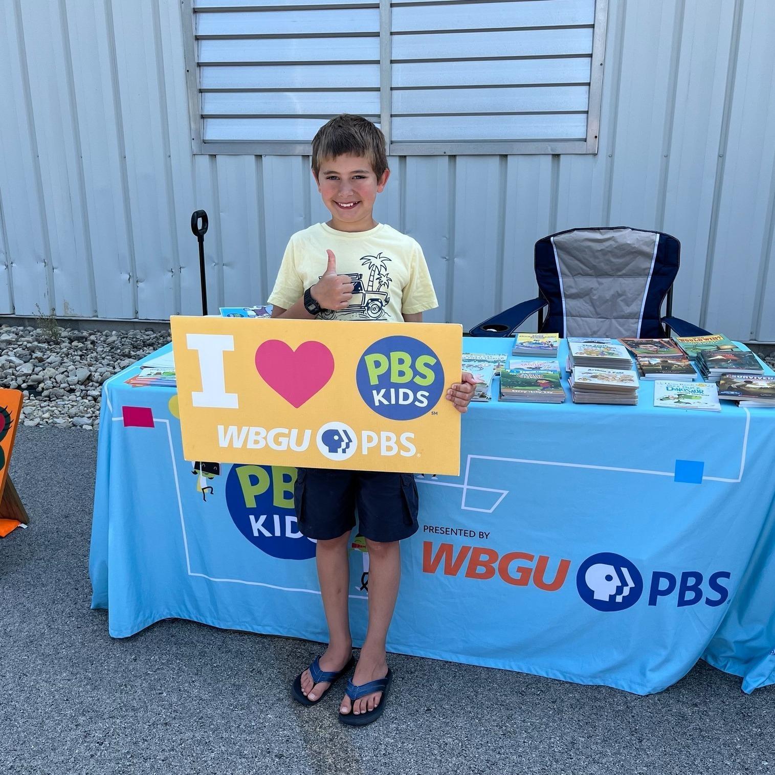 A smiling school aged boy gives a thumbs up and hold an I Love WBGU-PBS sign in front of WBGU's blue display table in the parking lot of the Findlay YMCA East Branch.