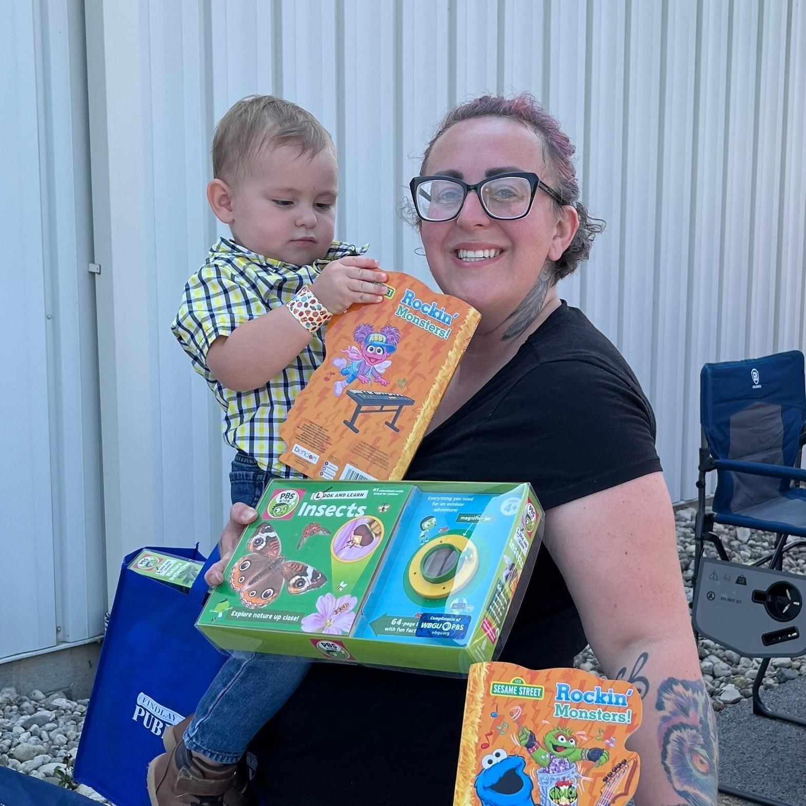 A smiling mother and toddler aged son display Sesame Street books and a PBS KIDS nature kit that they received from WBGU-PBS at the Findlay Summer Connect Event at the Findlay YMCA East Branch. The toddler is holding one of the books and attempting to open the book while he looks at it. The smiling mother is wearing glasses and looking directly at the camera. The mother is holding her son and standing outside the YMCA building in the event parking lot.