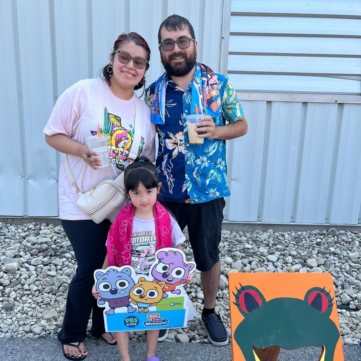 A smiling mother and father, wearing sunglasses and holding drinks stand behind their preschool aged daughter who is  holding a PBS KIDS Work It Out Wombats sign. They are standing to the left of a bean bag toss board with a painting of WBGU's mascot -- Ruby the Red-Eyed Tree Frog on it. 