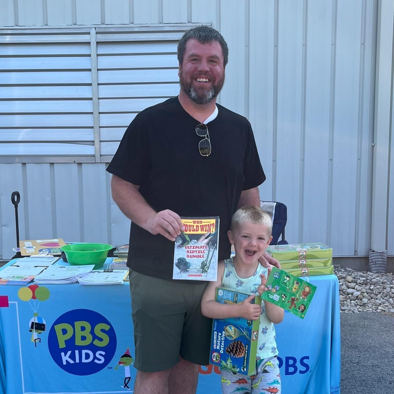 A smiling father with a smiling preschool aged son display the book, nature kit and PBS KIDS stickers that they received from WBGU-PBS at the Findlay Summer Connect Event at the Findlay YMCA East Branch. They are standing in front of a WBGU-PBS table with a light blue table cloth outside in the parking lot of the YMCA.