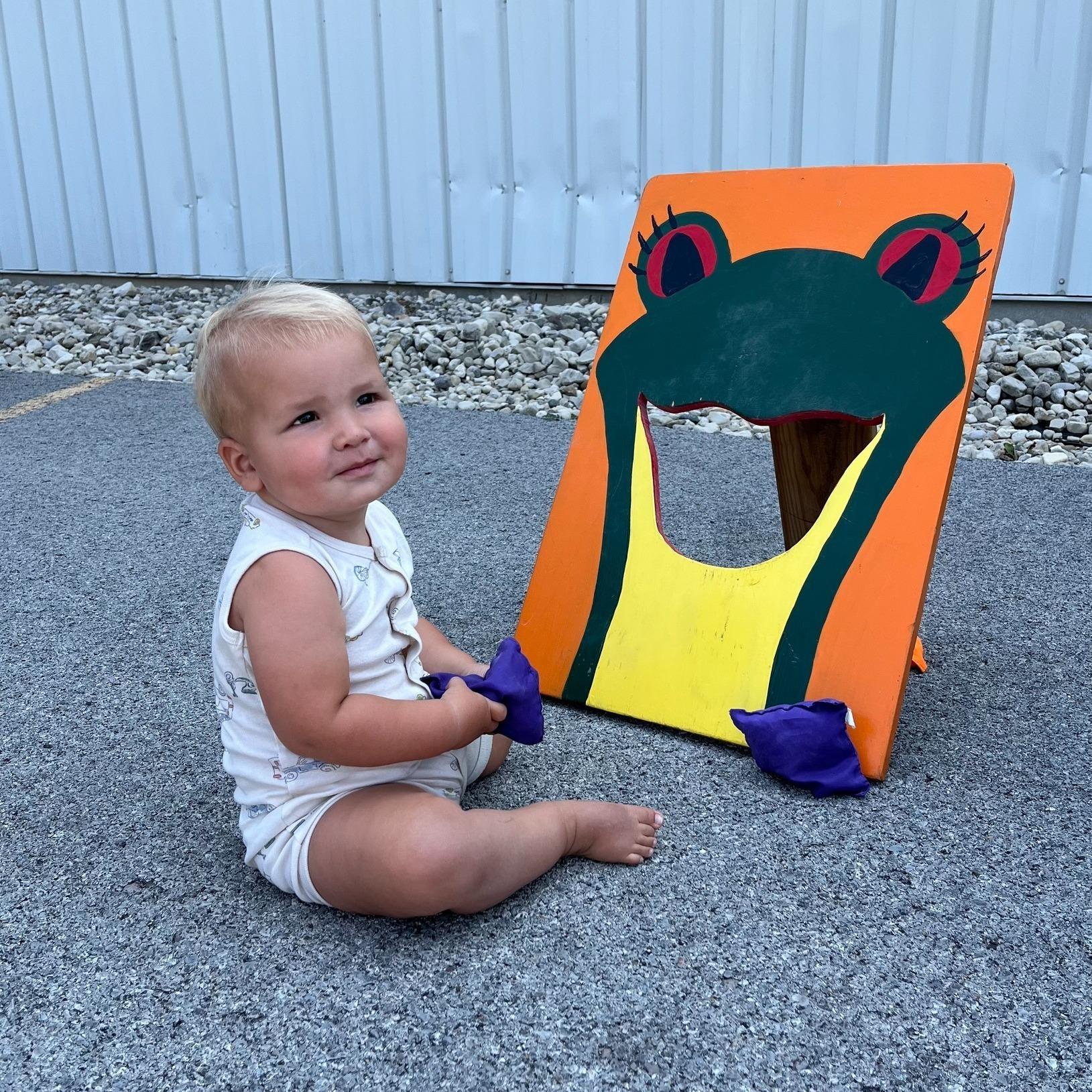 A grinning young toddler sits on the ground holding a purple bean bag near a wooden bean bag board that is painted to look like WBGU's mascot Ruby the Red Eyed Tree Frog with an open mouth. The toddler has blond hair and is wearing a white sleeveless romper.