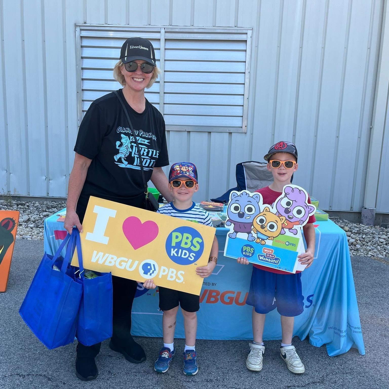 Smiling adult women with short blonde hair, wearing a black ball cap and sunglasses with a black t-shirt holds 2 blue tote bags and puts her arm around the shoulders of a child wearing a dark ball cap, orange sun glasses and a striped shirt and short. They are holding a yellow sign that say I heart (love) PBS KIDS and WBGU-PBS. Another slightly older boy is standing to the right of them holding a Work It Out Wombats foam sign. The boy is wearing a dark ball cap and orange sunglasses.